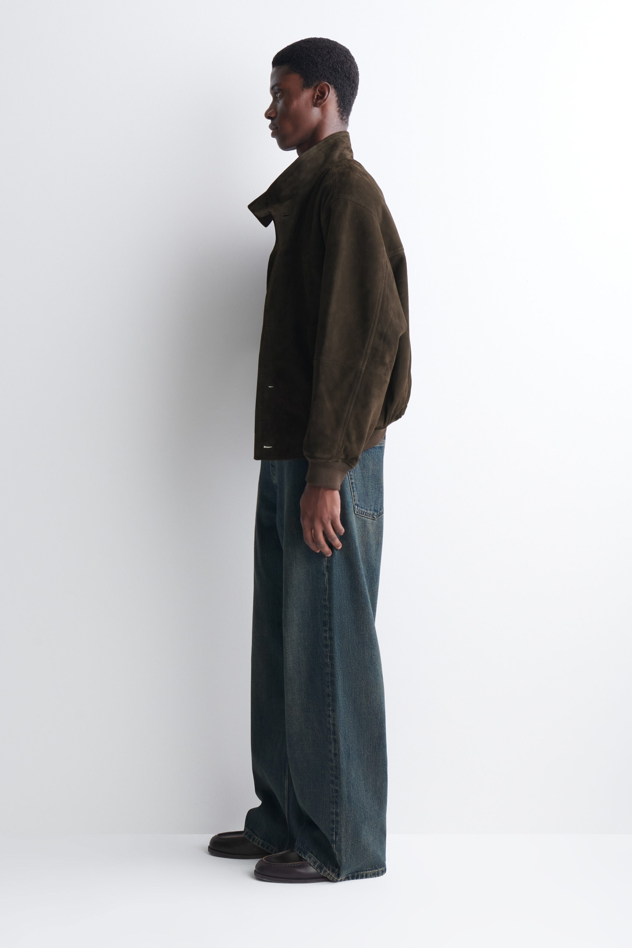 A dark brown suede jacket featuring a high collar, ribbed cuffs and a relaxed silhouette is being worn by the model, who is standing in profile. The jacket is styled with wide-leg, dark-wash blue jeans and dark leather shoes. A dark brown suede jacket featuring a high collar, ribbed cuffs and a relaxed silhouette is being worn by the model, who is standing in profile. The jacket is styled with wide-leg, dark-wash blue jeans and dark leather shoes.