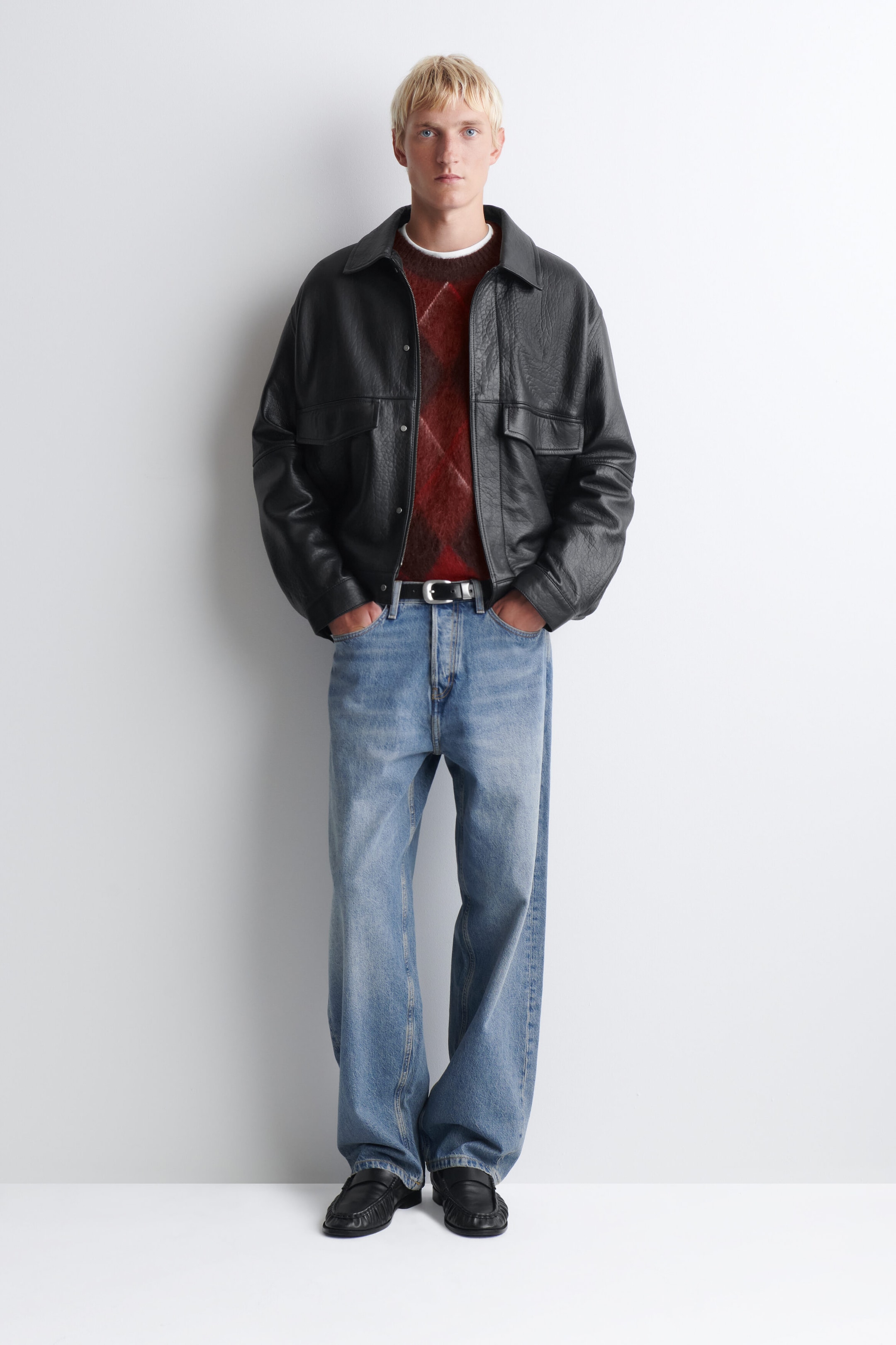 Eine schwarze Lederjacke ist von vorne zu sehen und zeichnet sich durch eine lockere Passform, einen spitzen Kragen, Druckknopfverschlüsse und grosse Brusttaschen mit Patte aus. Sie wird über einem rot und braun gemusterten Rundhalspullover und hell gewaschenen blauen Jeans mit weitem Bein getragen.