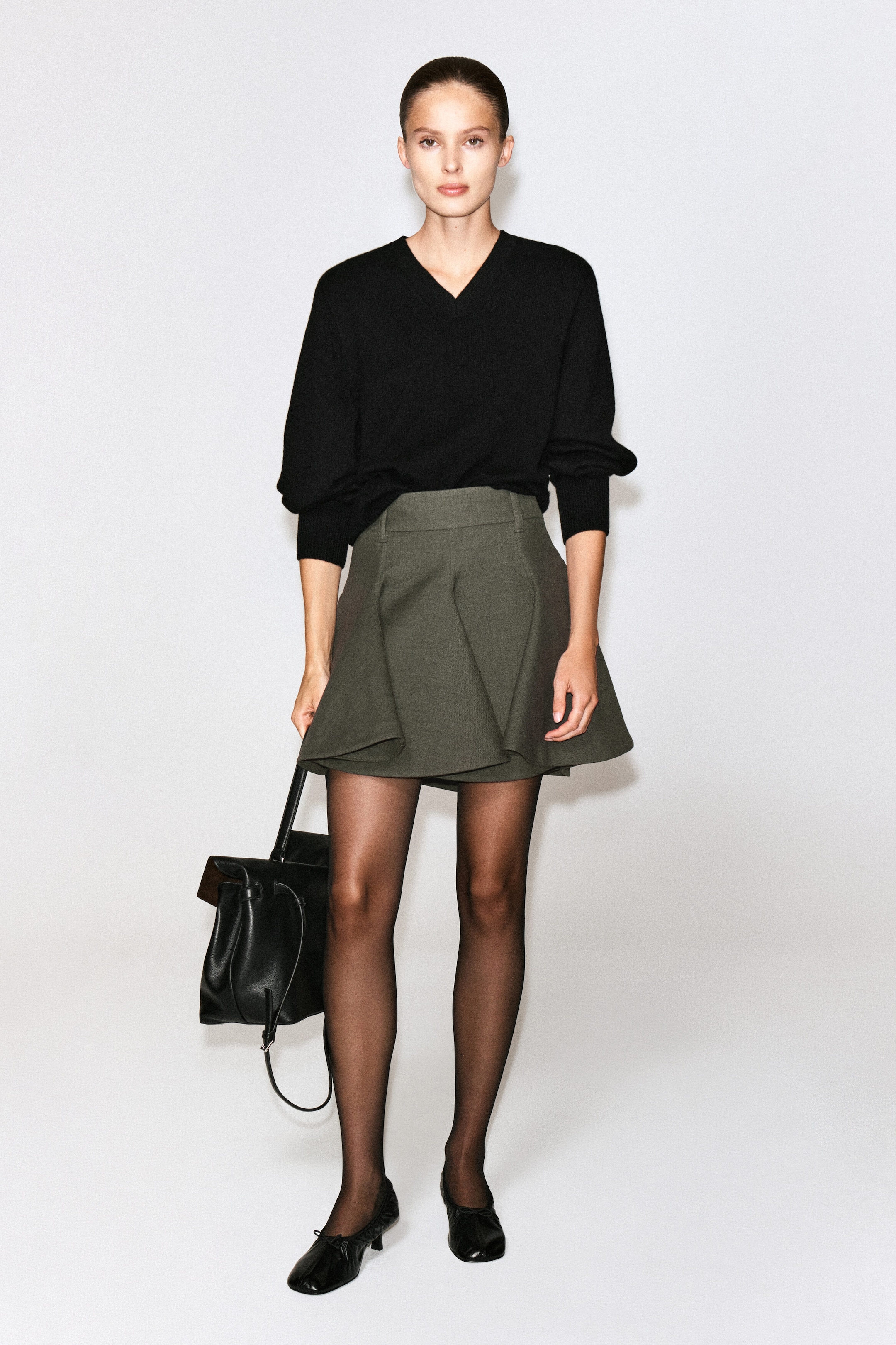 A dark olive green sculptural tailored wool mini skirt featuring structured folded pleats and a high waist is worn by the model. The skirt is styled with a tucked-in black V-neck sweater, sheer black tights, black pumps, and a black leather handbag held in one hand. A dark olive green sculptural tailored wool mini skirt featuring structured folded pleats and a high waist is worn by the model. The skirt is styled with a tucked-in black V-neck sweater, sheer black tights, black pumps, and a black leather handbag held in one hand.
