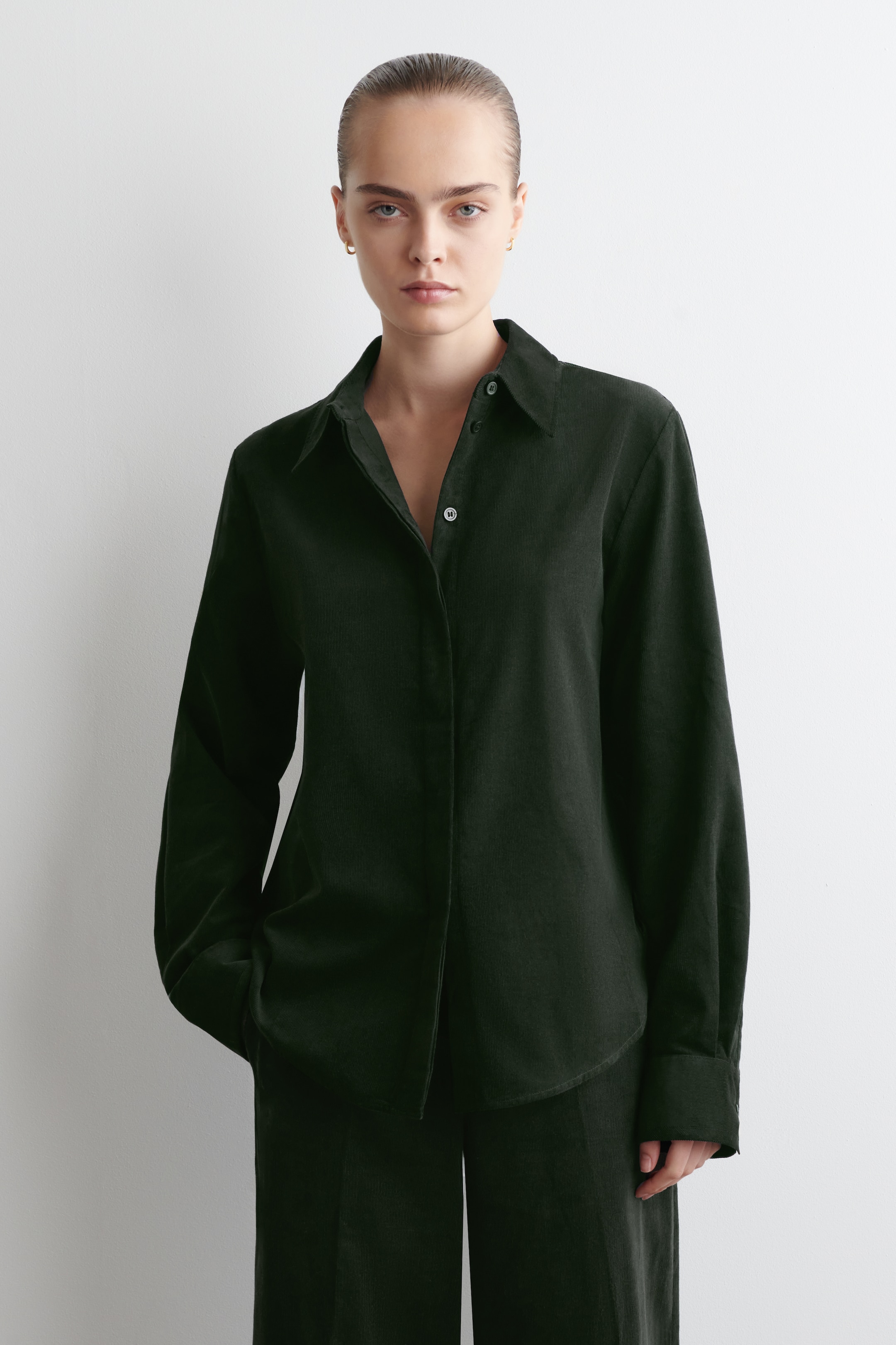Visualizza immagine più grande: Una camicia di velluto a coste verde scuro viene presentata, rivolta in avanti, con una vestibilità rilassata e maniche lunghe. È progettata con un classico colletto a punta e una chiusura frontale con bottoni, con diversi bottoni slacciati, ed è abbinata a pantaloni verde scuro coordinati.