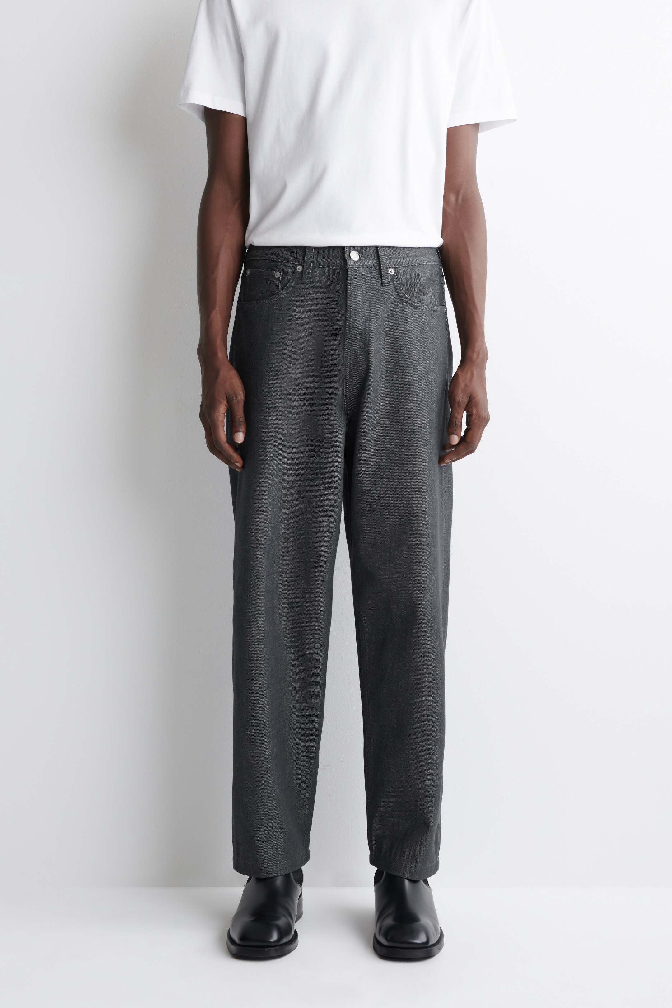 Agrandir l'image: Le mannequin porte un jean gris foncé à coupe décontractée avec une jambe droite et une fermeture à boutons visible à la taille mi-haute, ainsi qu'un t-shirt blanc et des chaussures noires.