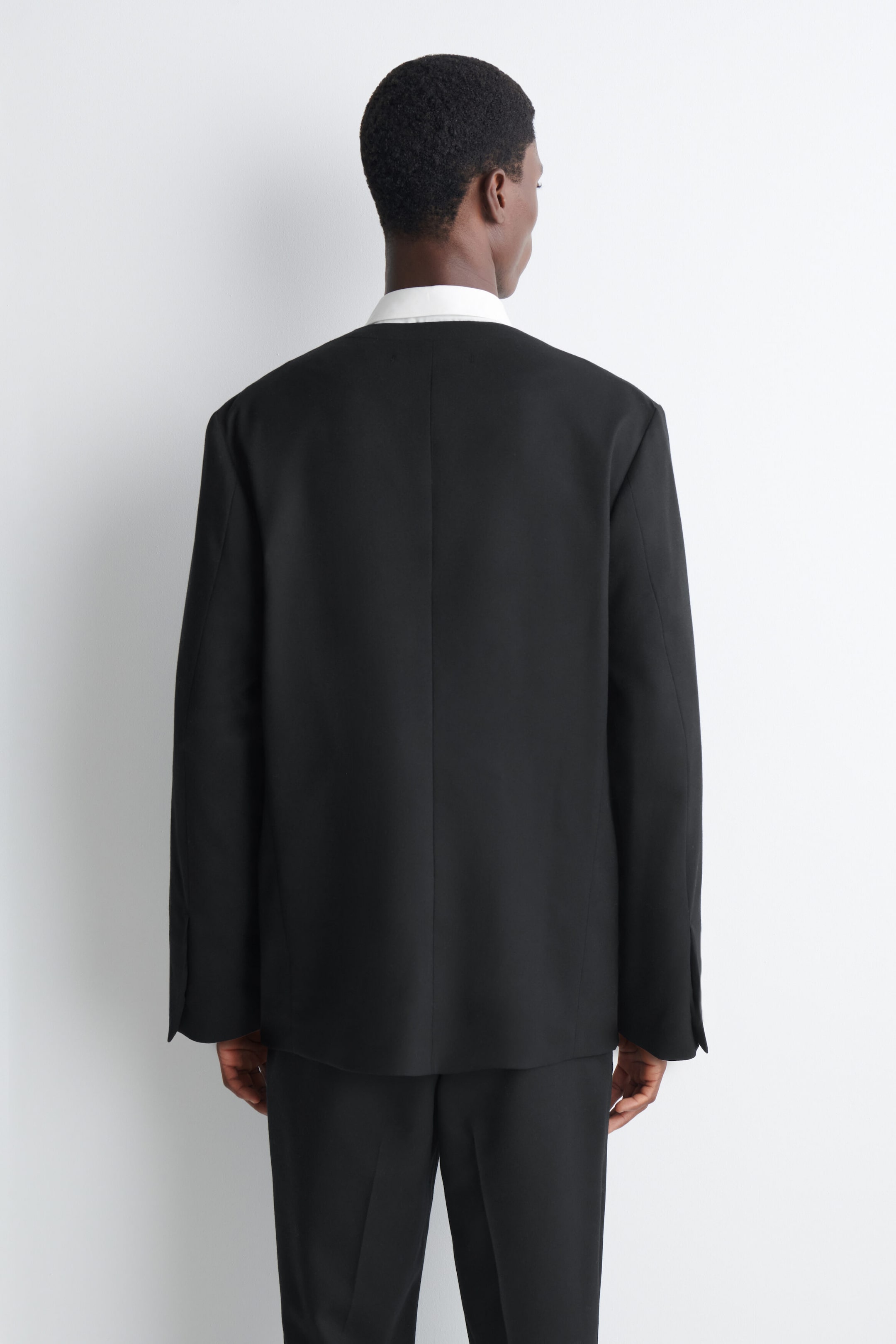The model is wearing a black collarless blazer, shown from the back, featuring a structured, minimalist design with a centre back seam. The jacket is layered over a white collared shirt and paired with matching black trousers. The model is wearing a black collarless blazer, shown from the back, featuring a structured, minimalist design with a centre back seam. The jacket is layered over a white collared shirt and paired with matching black trousers.