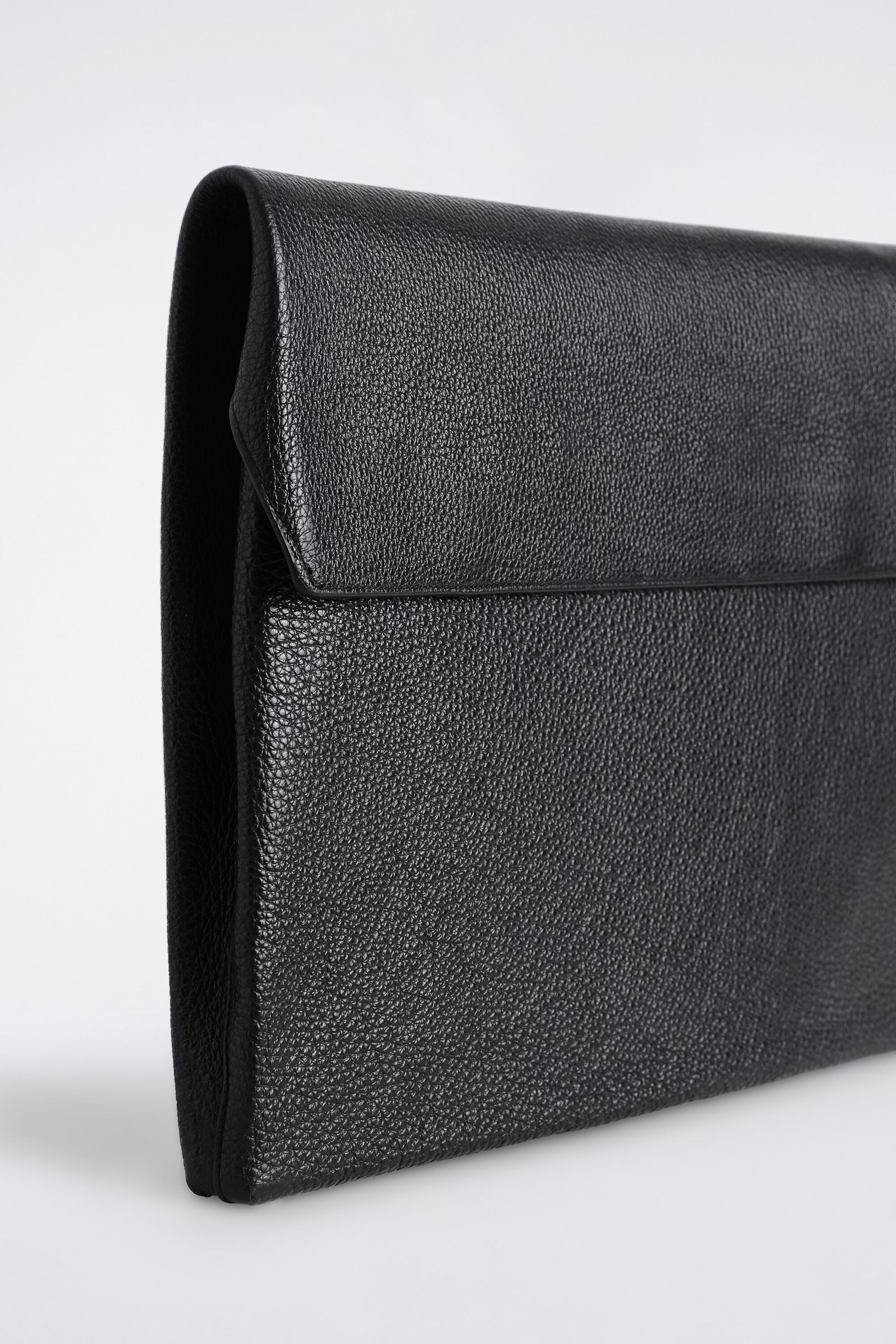 The black pebbled leather envelope laptop sleeve is shown at a three-quarter angle, highlighting its textured surface, fold-over top flap, and structured side profile. The black pebbled leather envelope laptop sleeve is shown at a three-quarter angle, highlighting its textured surface, fold-over top flap, and structured side profile.