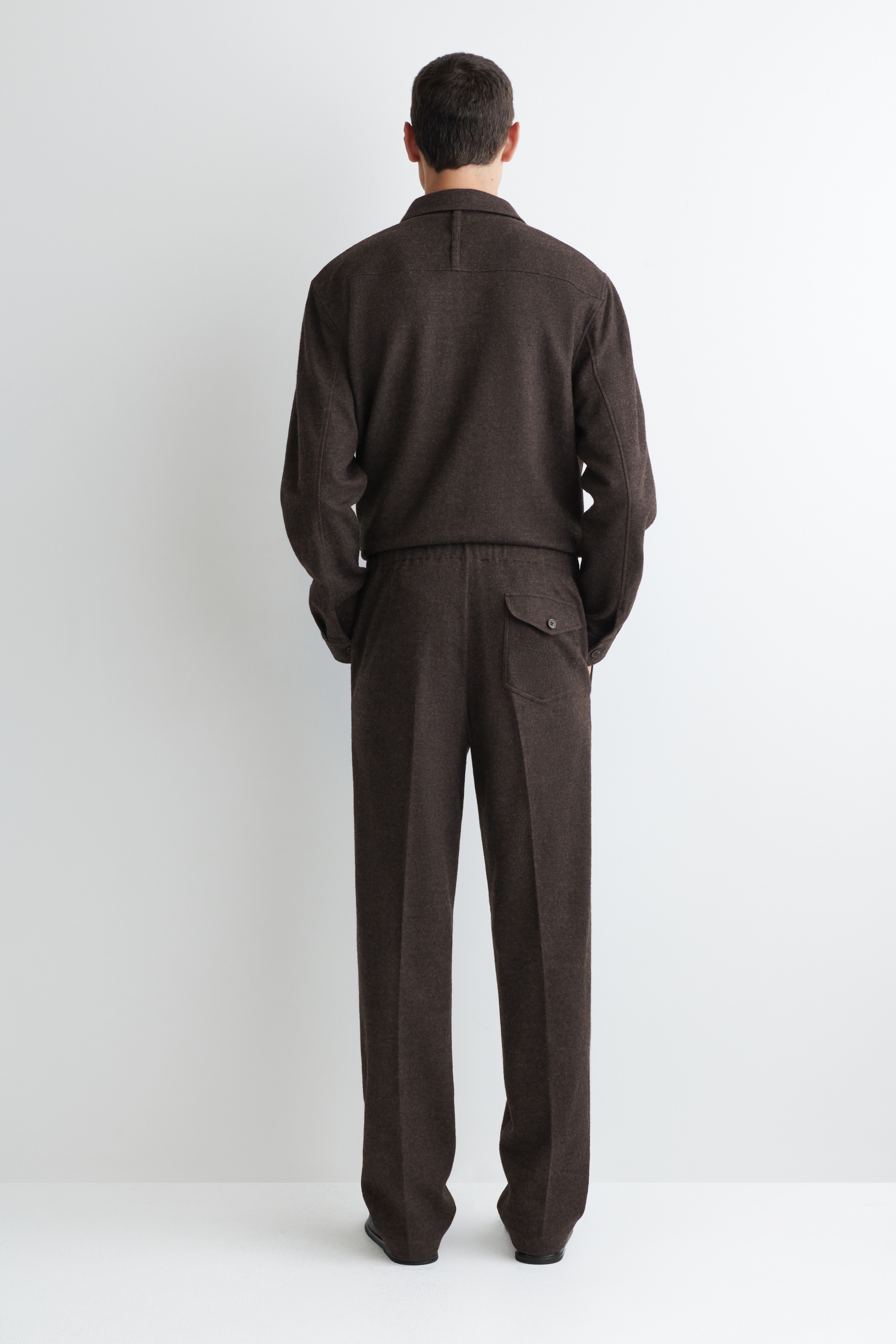 Visualizza immagine più grande: Un completo abbinato di giacca e pantaloni marrone e testurizzato viene presentato dal retro. La giacca presenta un colletto in stile camicia, un orlo elasticizzato e un piccolo dettaglio a passante sulla parte superiore della schiena. I pantaloni mostrano una vestibilità rilassata e dritta, una cintura elasticizzata e una singola tasca con patta e bottone sul retro destro.