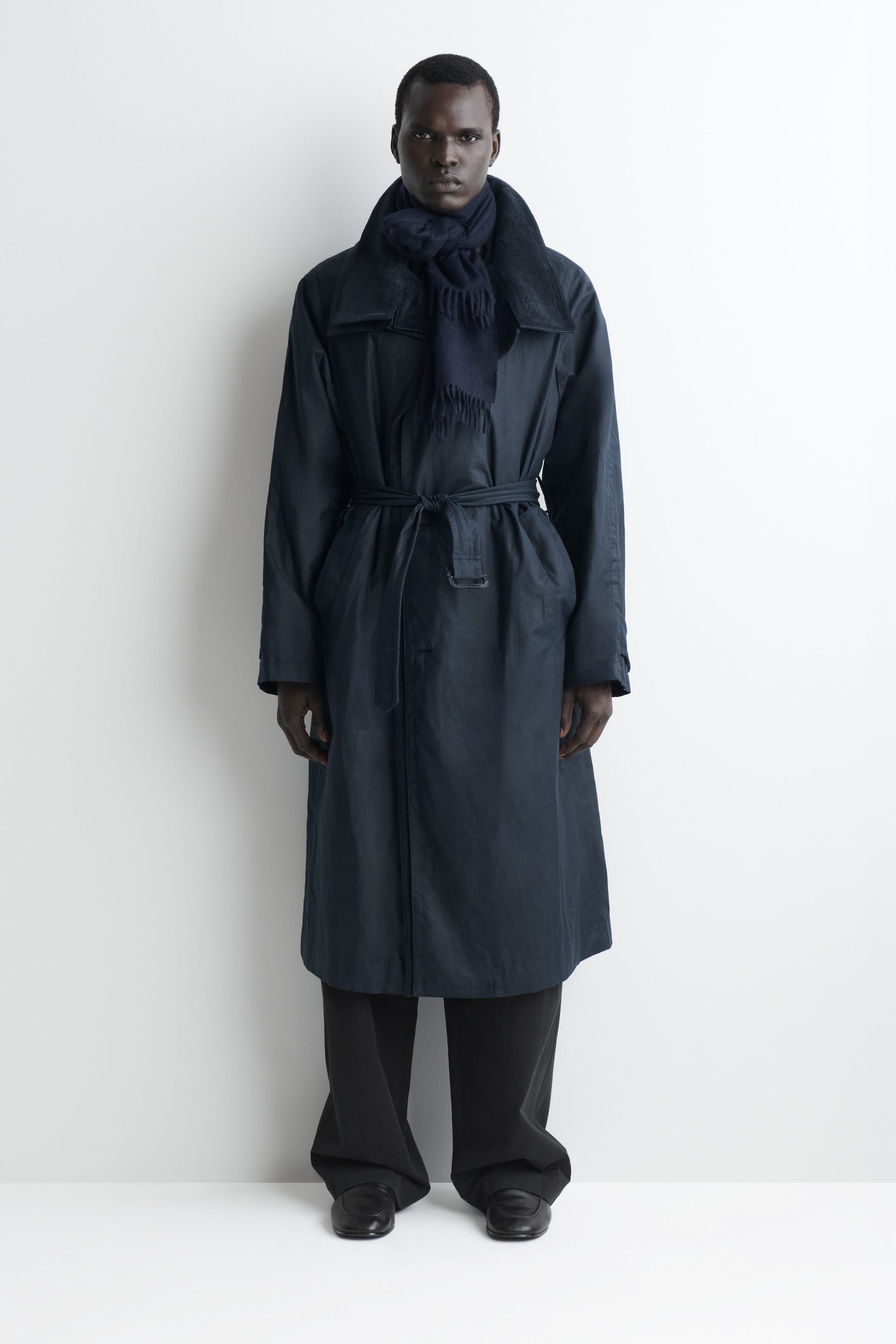 A navy blue fringed cashmere scarf is being worn wrapped around the neck of a model facing forward, with its fine-knit texture and delicate fringed ends hanging down the chest over a belted coat. A navy blue fringed cashmere scarf is being worn wrapped around the neck of a model facing forward, with its fine-knit texture and delicate fringed ends hanging down the chest over a belted coat.