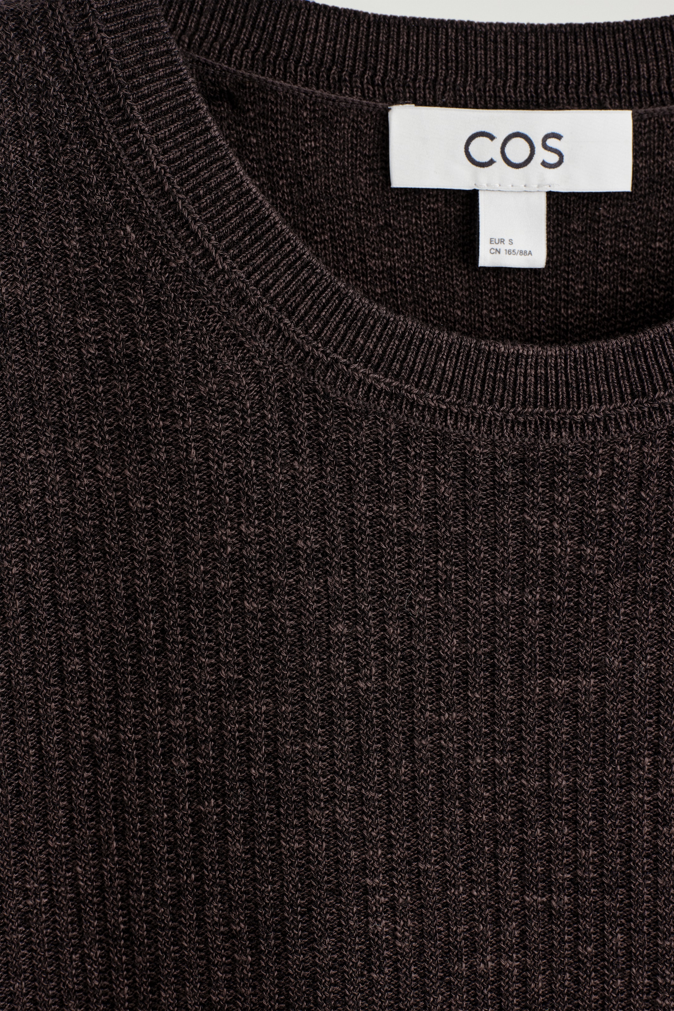 View larger image: This dark brown garment is featuring a close-up of its ribbed knit pattern, including a ribbed round neckline, and a white brand label with 'COS' text is visible at the neck.