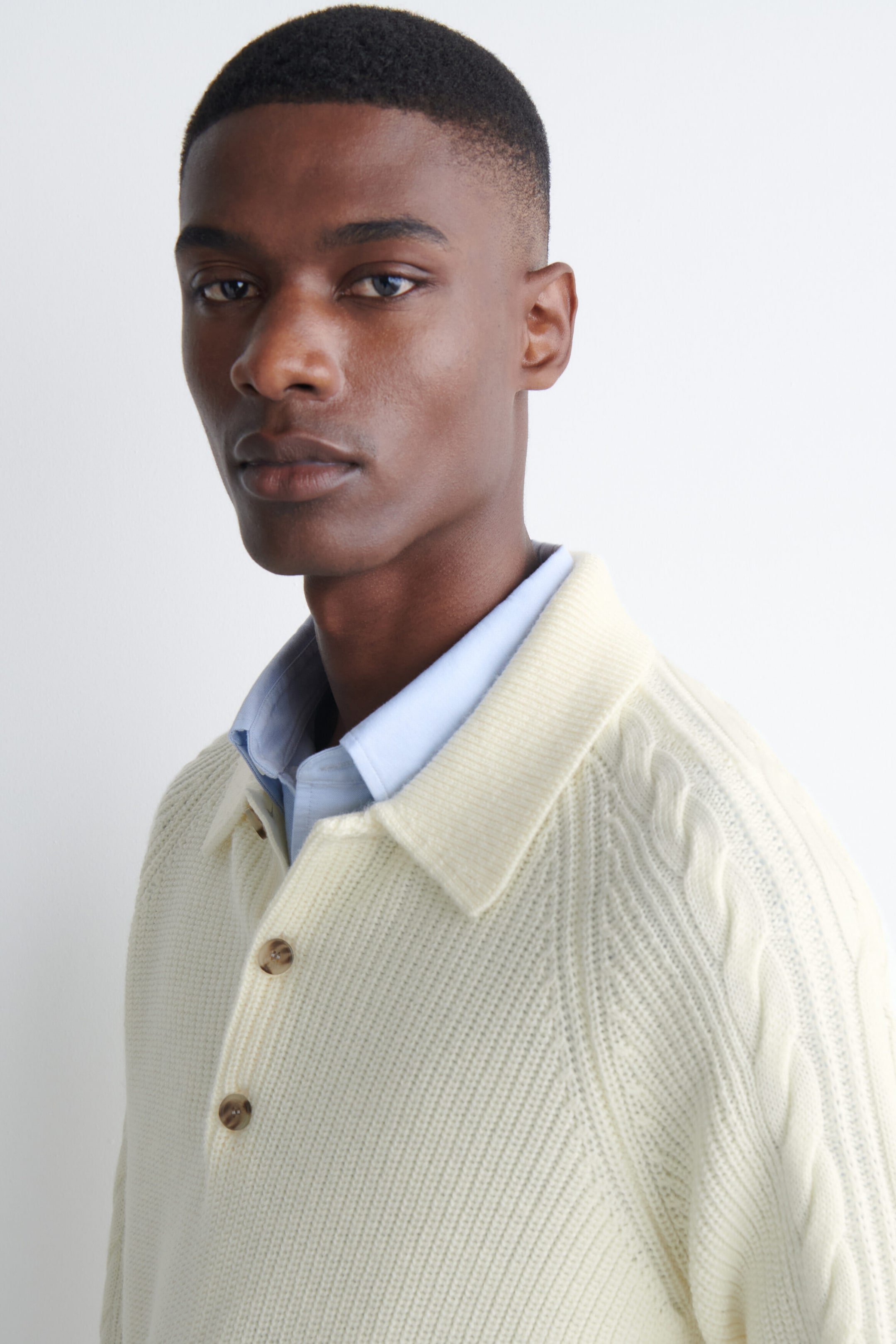 A cream-colored chunky knit polo sweater with a collar, button placket, and cable-knit sleeve details is worn by the model. The polo is layered over a light blue collared shirt. A cream-colored chunky knit polo sweater with a collar, button placket, and cable-knit sleeve details is worn by the model. The polo is layered over a light blue collared shirt.