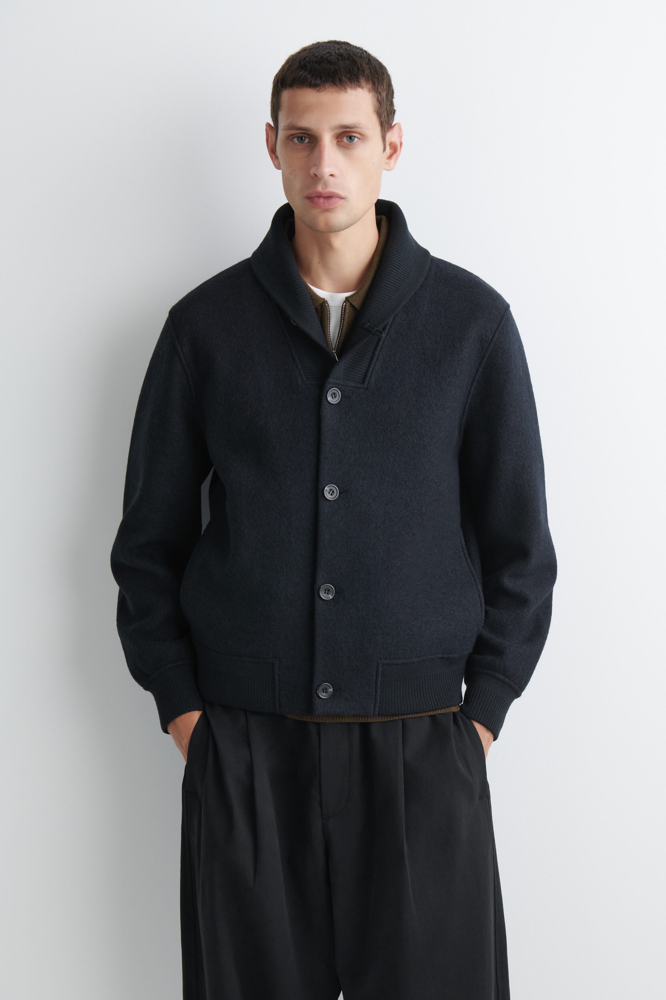 A dark navy boiled-wool shawl-collar jacket is shown from the front, featuring a four-button closure, ribbed cuffs, and a ribbed hem. The jacket is layered over a brown zippered shirt and a white T-shirt, highlighting its relaxed, structured fit. A dark navy boiled-wool shawl-collar jacket is shown from the front, featuring a four-button closure, ribbed cuffs, and a ribbed hem. The jacket is layered over a brown zippered shirt and a white T-shirt, highlighting its relaxed, structured fit.