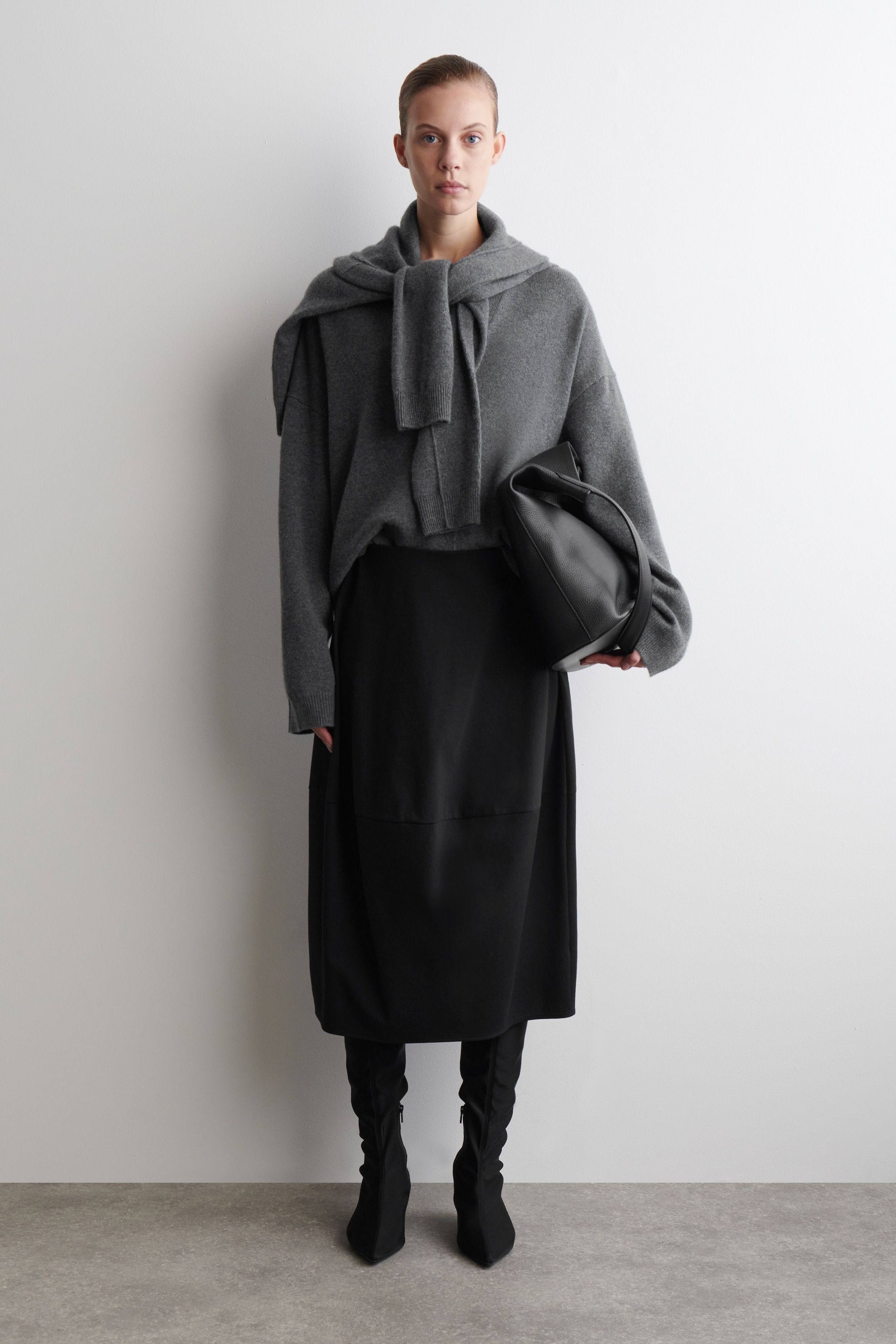 Agrandir l'image: Une tenue complète est présentée de face, avec un pull en maille gris à coupe décontractée et manches longues, ainsi qu'un pull en maille gris assorti drapé et noué à l'avant. L'ensemble comprend une jupe noire de longueur midi avec des détails de panneaux, des bottes noires à bout pointu avec des tiges froncées, et un sac à main texturé noir tenu sur le côté.