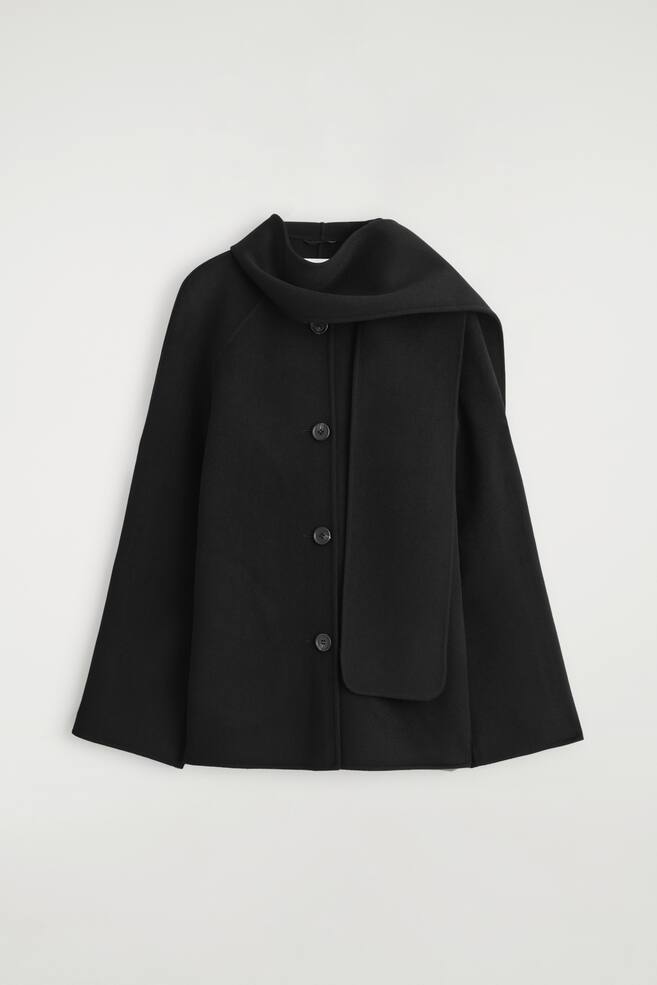 A black wool-blend jacket is shown facing FRONT, featuring an integrated high collar that extends into a long, wide scarf draping over the shoulders and down the front. It is designed with a relaxed A-line silhouette, wide raglan sleeves, and a front button closure with three visible buttons. - 1