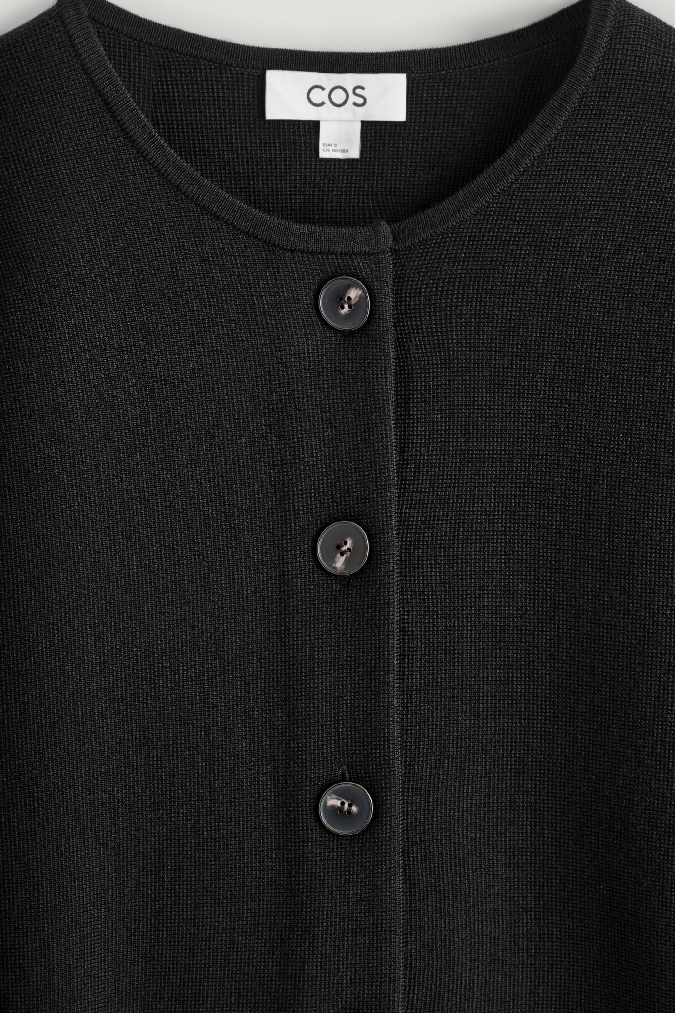 Größeres Bild ansehen: Eine schwarze Strickjacke ist nach vorne gerichtet und betont ihren runden Halsausschnitt. Die Strickjacke verfügt über eine Knopfleiste mit drei dunklen, marmorierten Knöpfen, und ihr Stoff zeigt eine feine, dichte Strickstruktur.