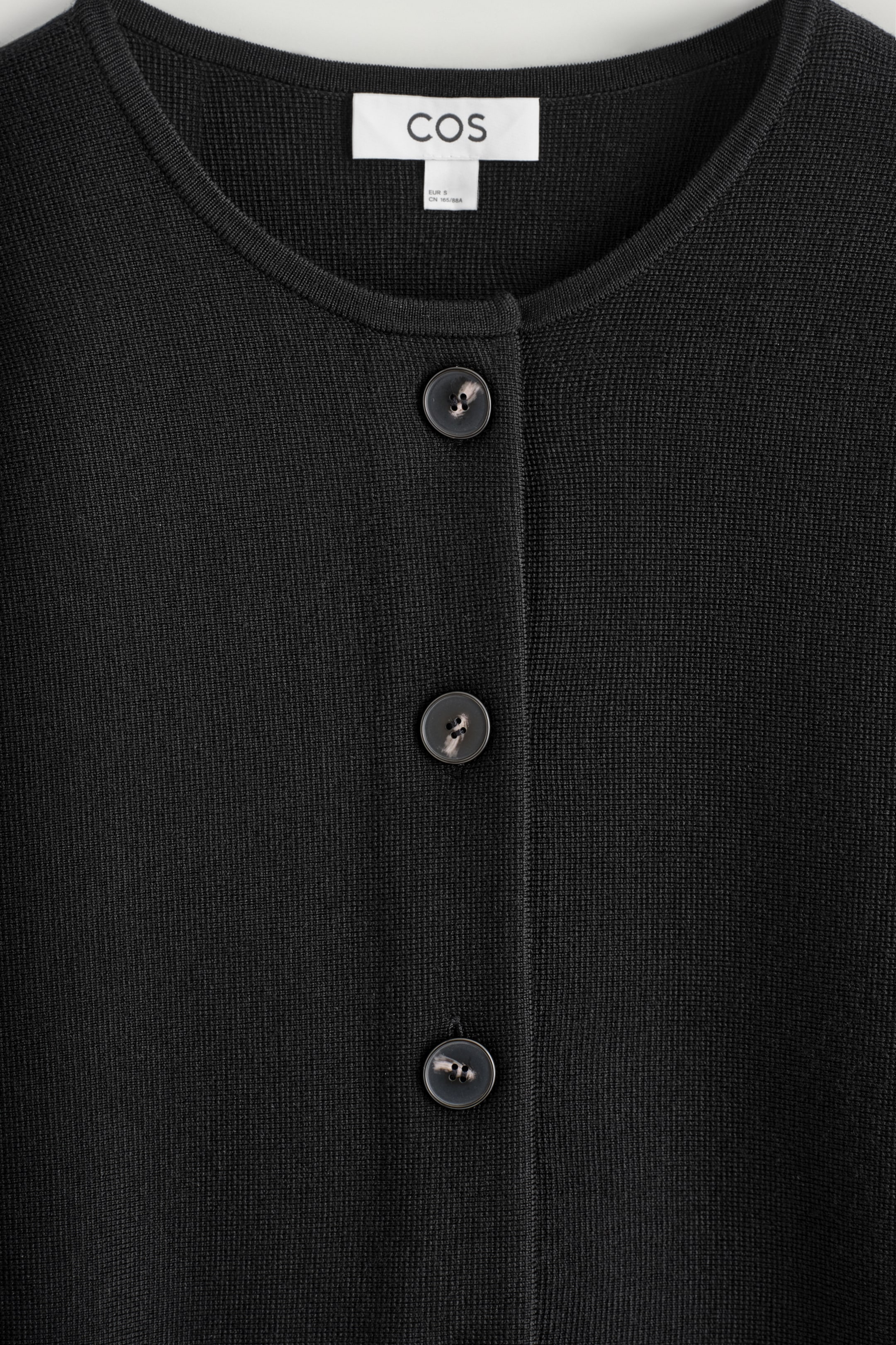 Agrandir l'image: Un cardigan en tricot noir est positionné face à l'avant, mettant en valeur son encolure ronde. Le cardigan présente une patte de boutonnage avec trois boutons sombres et marbrés, et son tissu exhibe une texture de tricot fine et dense.