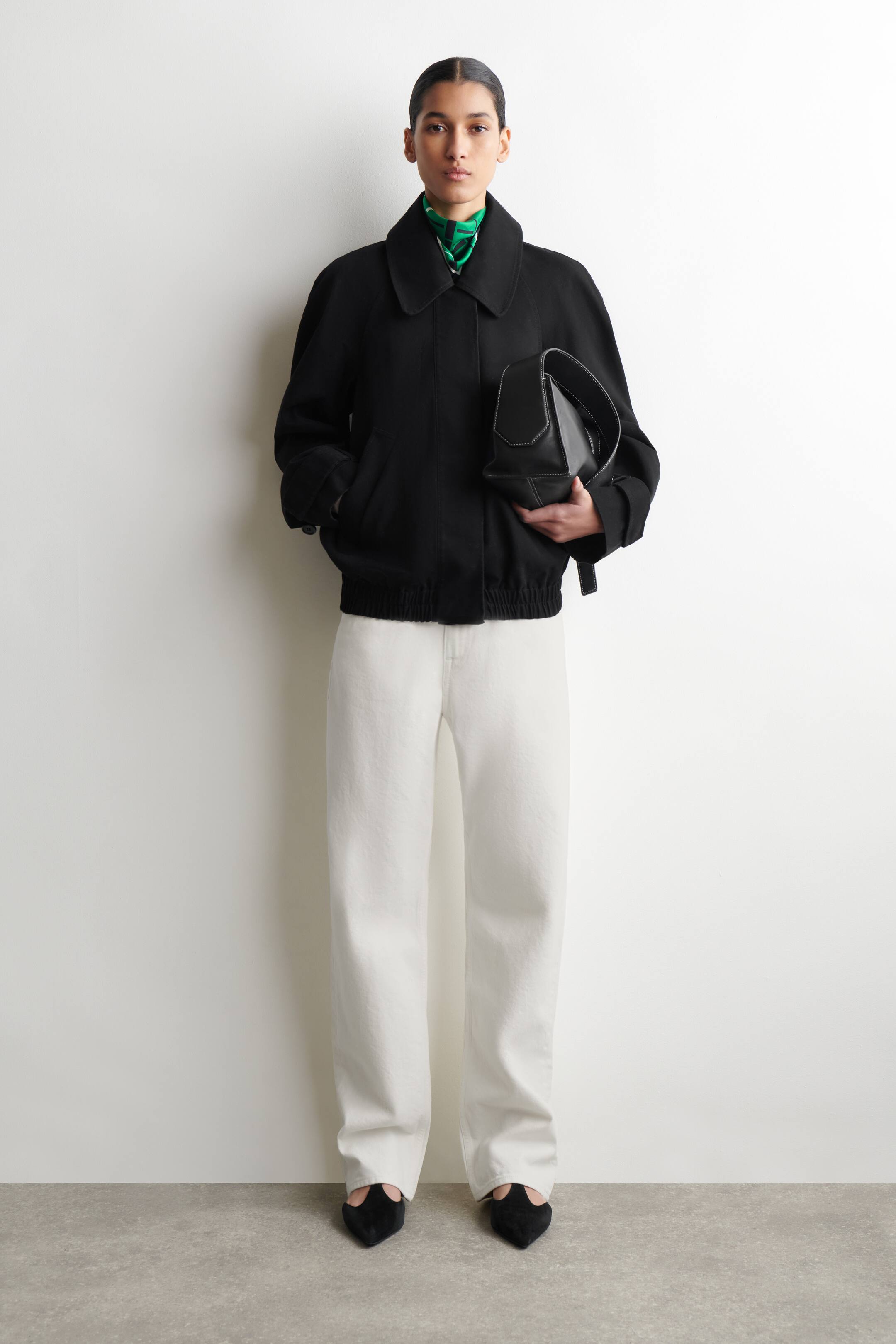 Agrandir l'image: Une veste bomber noire est présentée de face, avec un col rond et évasé et des manches longues dotées de poignets élastiques. La veste est conçue avec une coupe décontractée et un ourlet élastique, tandis que des poches latérales sont visibles. Un foulard à motifs verts se glisse sous le col.