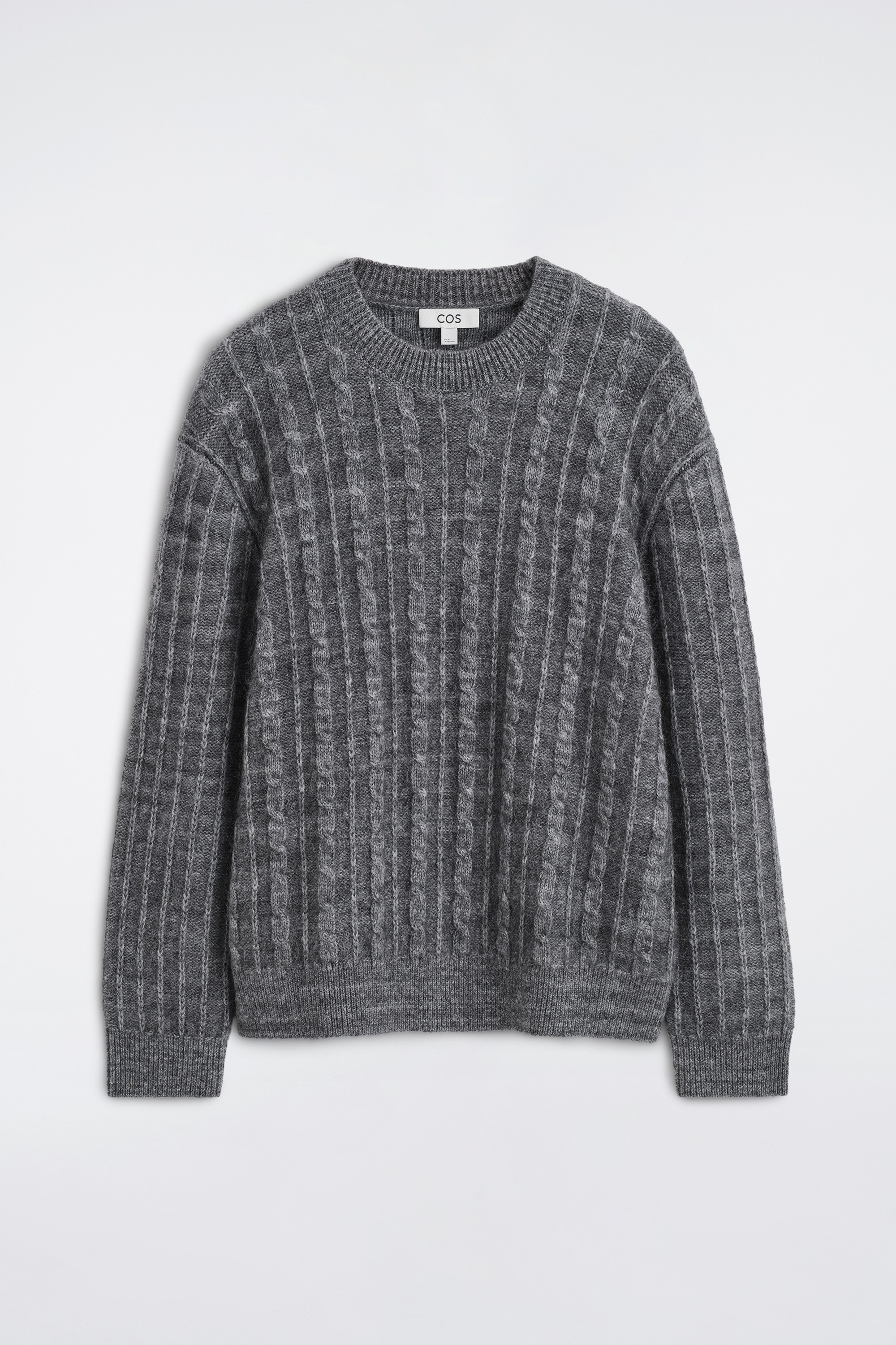 Un maglione grigio lavorato a trecce, con una finitura testurizzata e morbida, è indossato dalla modella. Il maglione presenta un girocollo e polsini a costine ed è infilato in pantaloni neri plissettati, fermati da una cintura nera.
