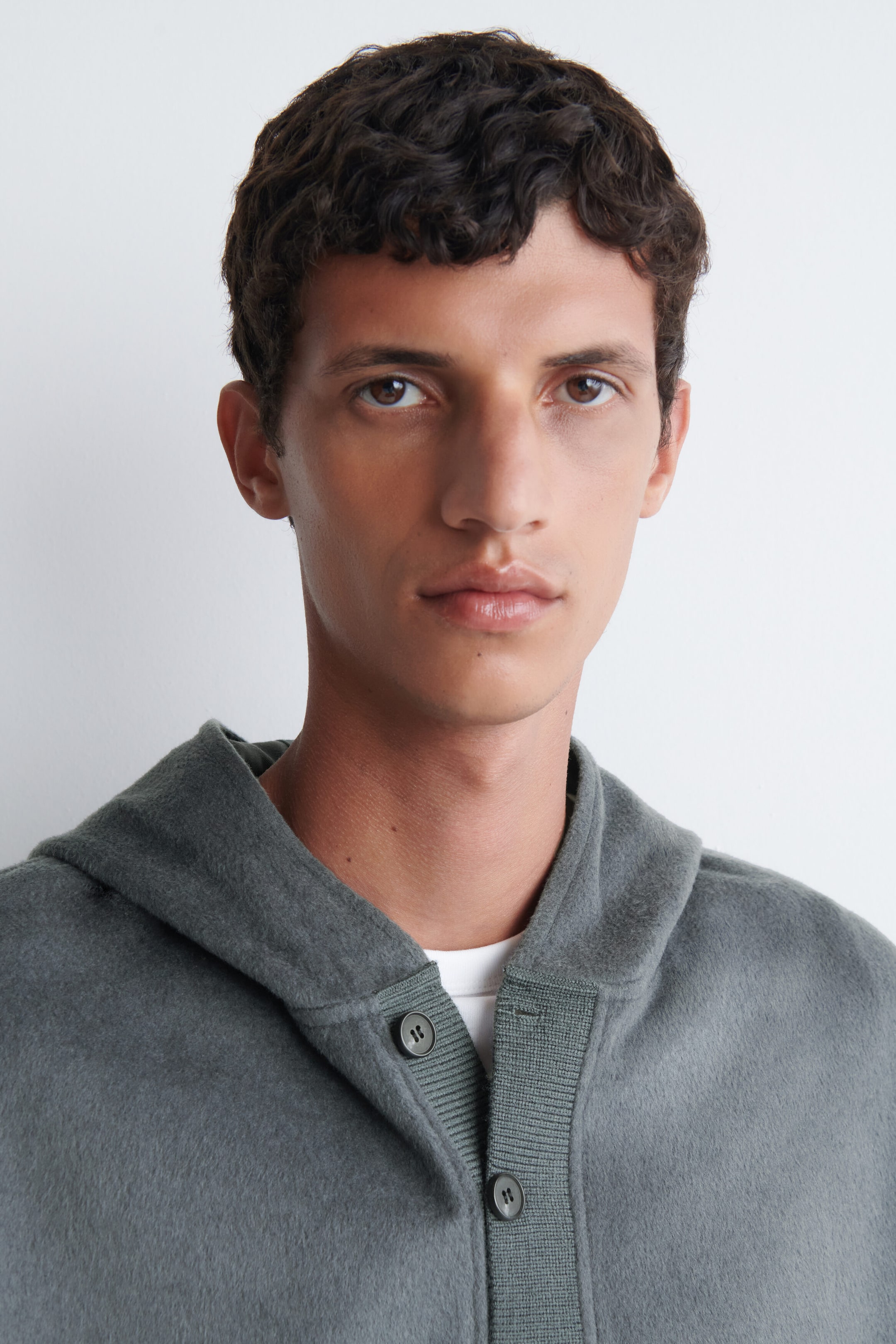 La mannequin porte une veste à capuchon gris-vert avec une texture brossée douce et une patte de boutonnage côtelée. La veste est superposée à un t-shirt blanc à col rond. La mannequin porte une veste à capuchon gris-vert avec une texture brossée douce et une patte de boutonnage côtelée. La veste est superposée à un t-shirt blanc à col rond.