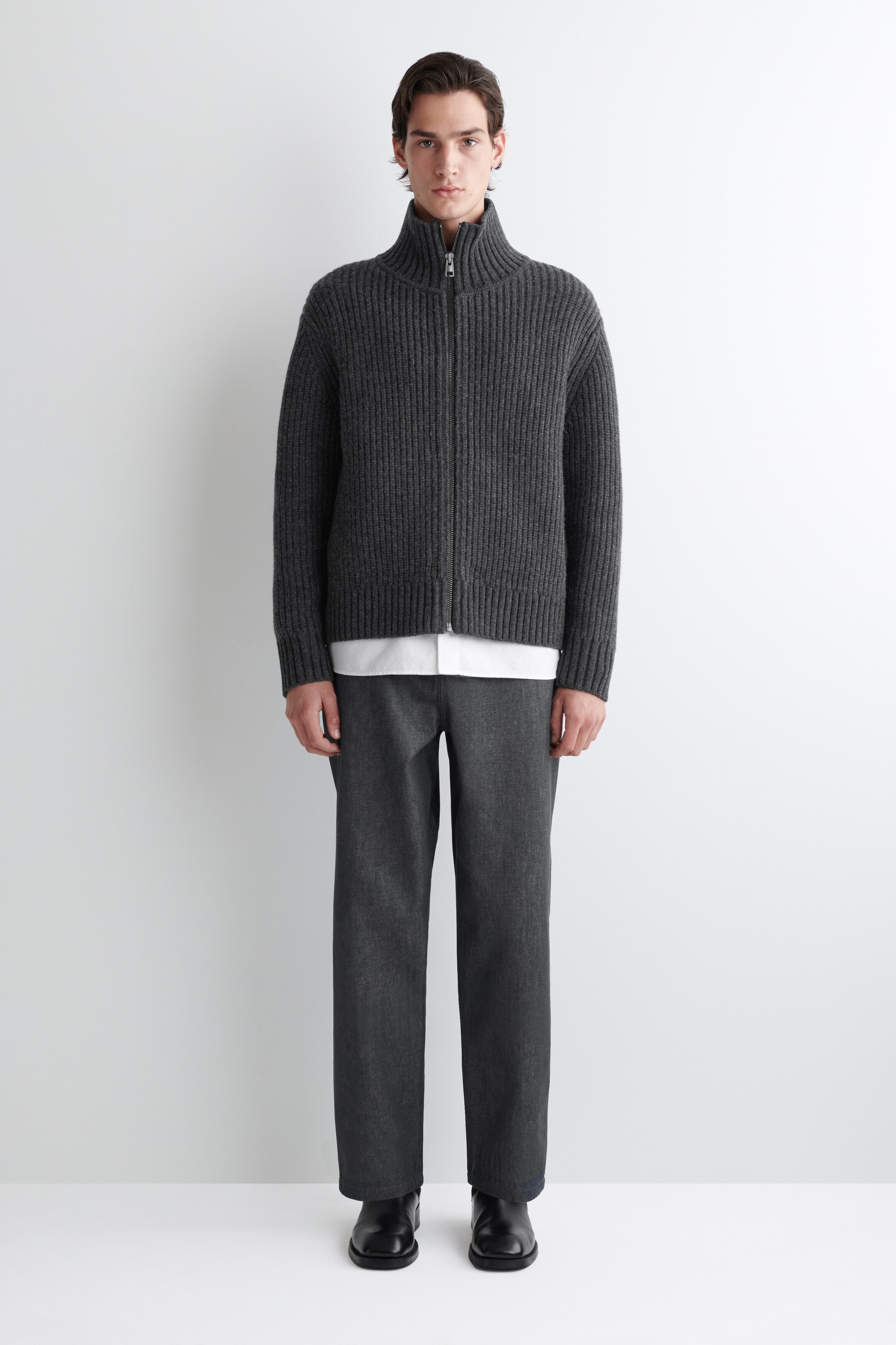 Agrandir l'image: Un cardigan à fermeture éclair complète, de couleur gris foncé et côtelé, est présenté face avant, avec un col montant et une fermeture éclair argentée sur le devant. Il est superposé à une chemise blanche, qui est visible sous l'ourlet du cardigan.