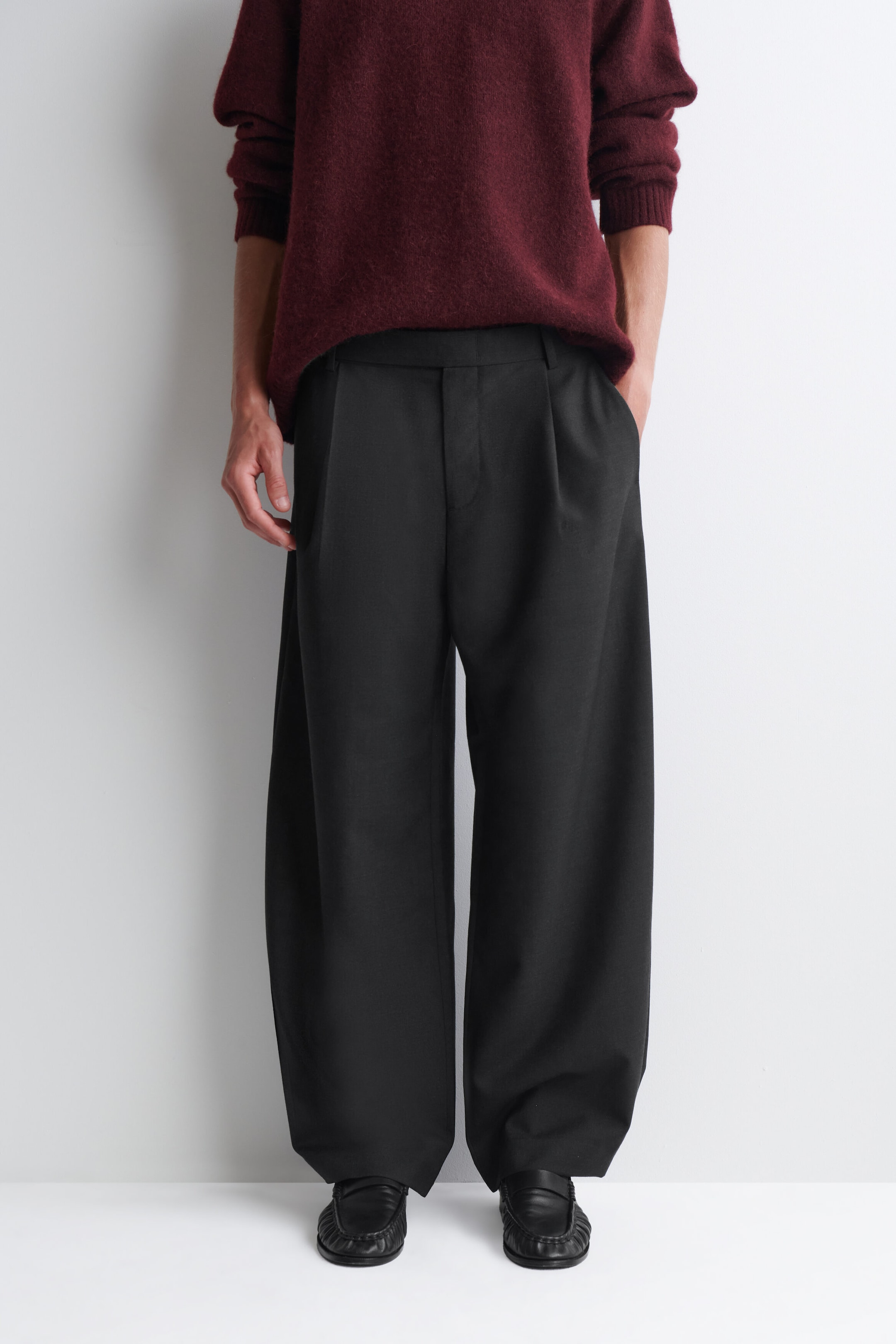 The black belt-detail wool relaxed-fit barrel-leg trousers are shown from the front, showcasing their wide, curved silhouette and front pleats. The relaxed-fit trousers drape over black leather loafers, with a burgundy knit jumper partially tucked into the waistband. The black belt-detail wool relaxed-fit barrel-leg trousers are shown from the front, showcasing their wide, curved silhouette and front pleats. The relaxed-fit trousers drape over black leather loafers, with a burgundy knit jumper partially tucked into the waistband.