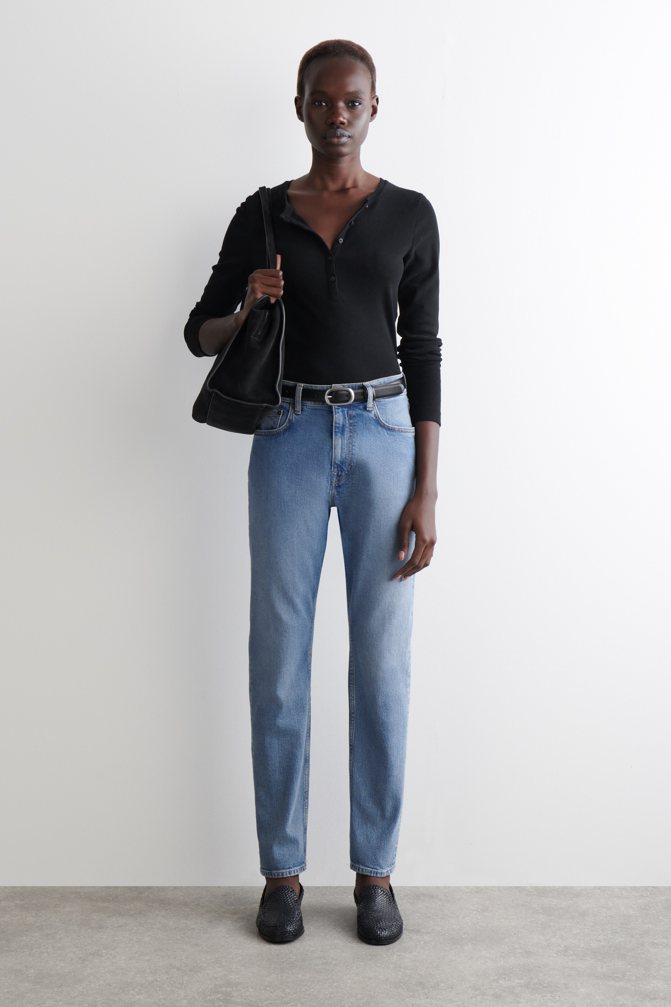 Agrandir l'image: Un haut Henley noir à manches longues est présenté, avec une patte de boutonnage. Il est porté avec un jean bleu à coupe droite et délavage clair, une ceinture noire, des mocassins noirs tressés et un sac à bandoulière noir.