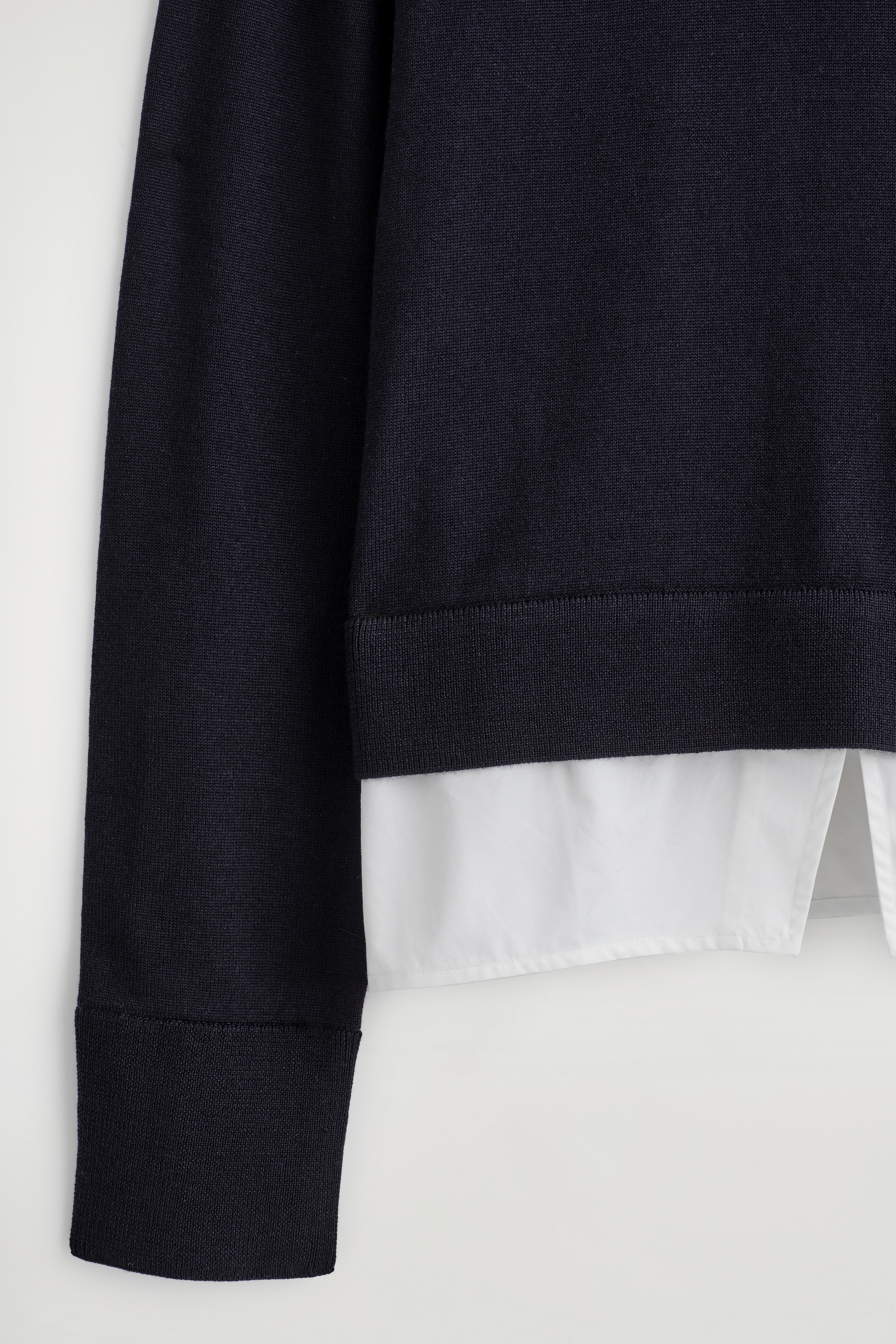 Visualizza immagine più grande: Un maglione a maglia fine di colore blu scuro, mostrato dal davanti, presenta un orlo a coste e un polsino a coste sulla manica sinistra, con un pannello di camicia in cotone bianco visibilmente stratificato sotto l'orlo, creando un effetto due in uno.
