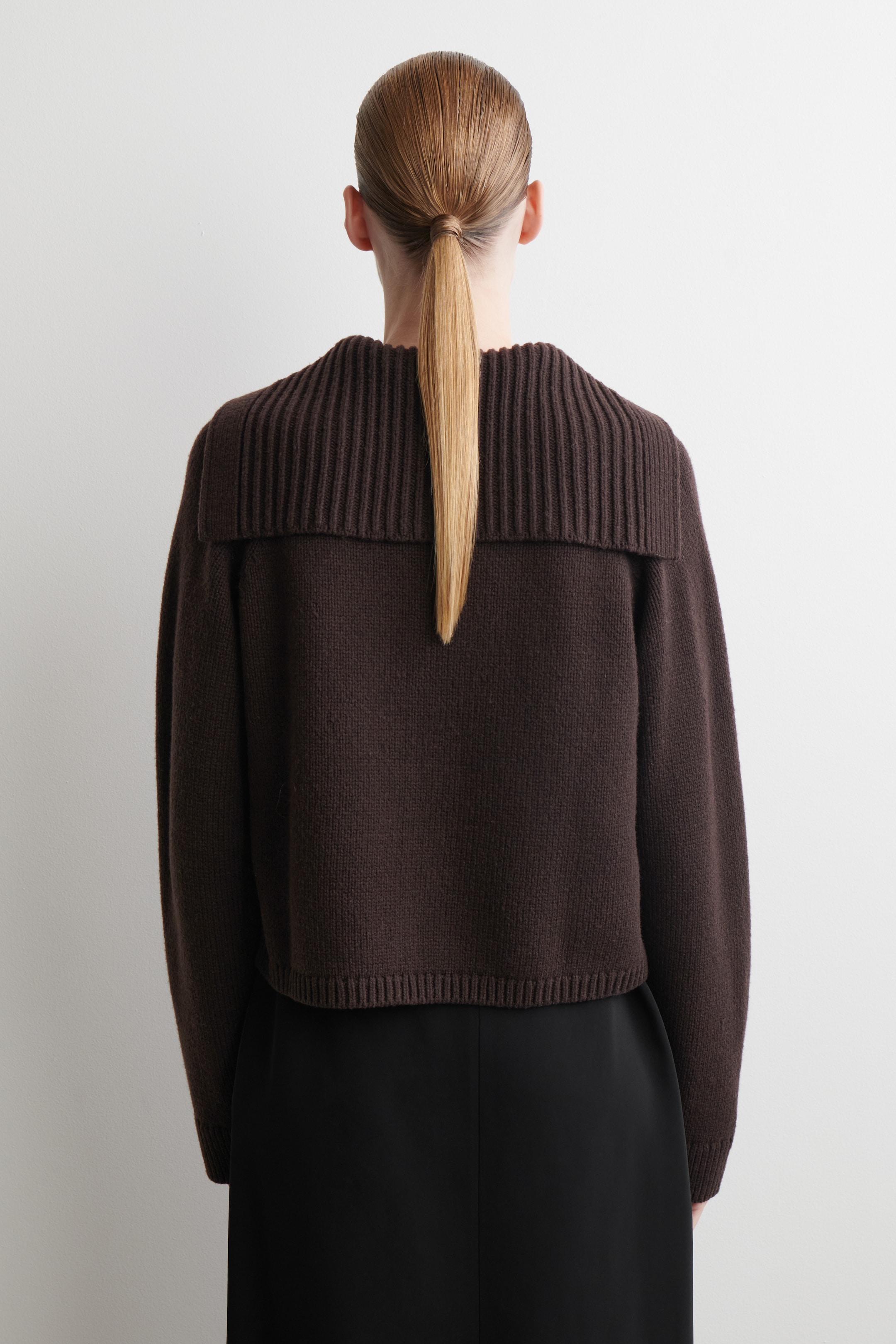 Visualizza immagine più grande: Un maglione marrone scuro viene visto da dietro. Presenta un ampio collo a coste ripiegato e un orlo a coste che termina appena sopra una gonna nera, mettendo in evidenza il suo design corto.