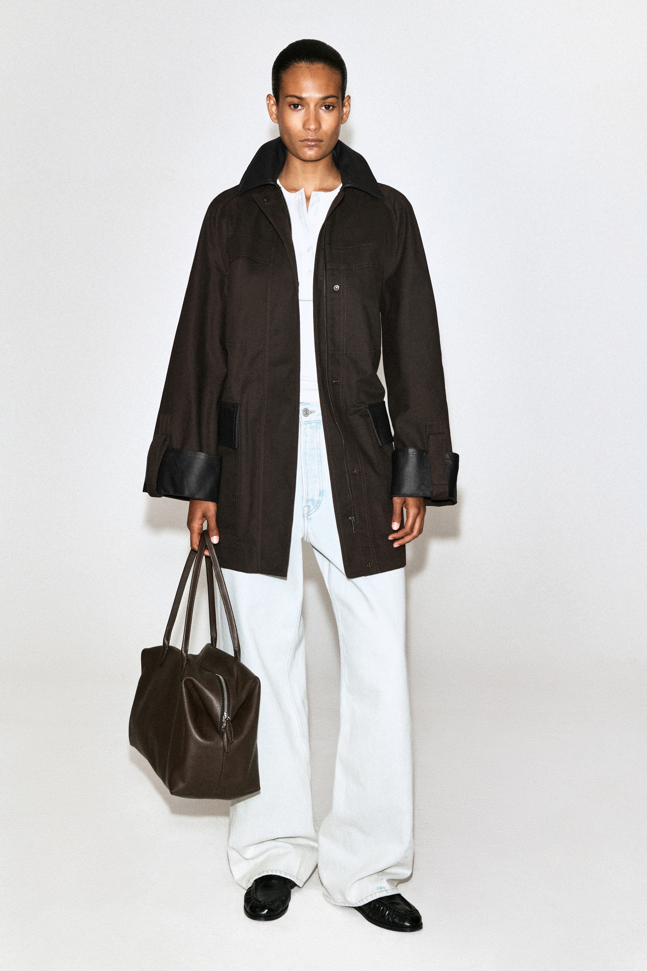 Agrandir l'image: Un manteau de campagne marron foncé, doté d’un col noir contrastant, de poignets retroussés révélant des détails en cuir noir grainé, de deux poches à rabat inférieures et d’une fermeture frontale à pression, est porté ouvert par le mannequin sur un sous-pull blanc.
