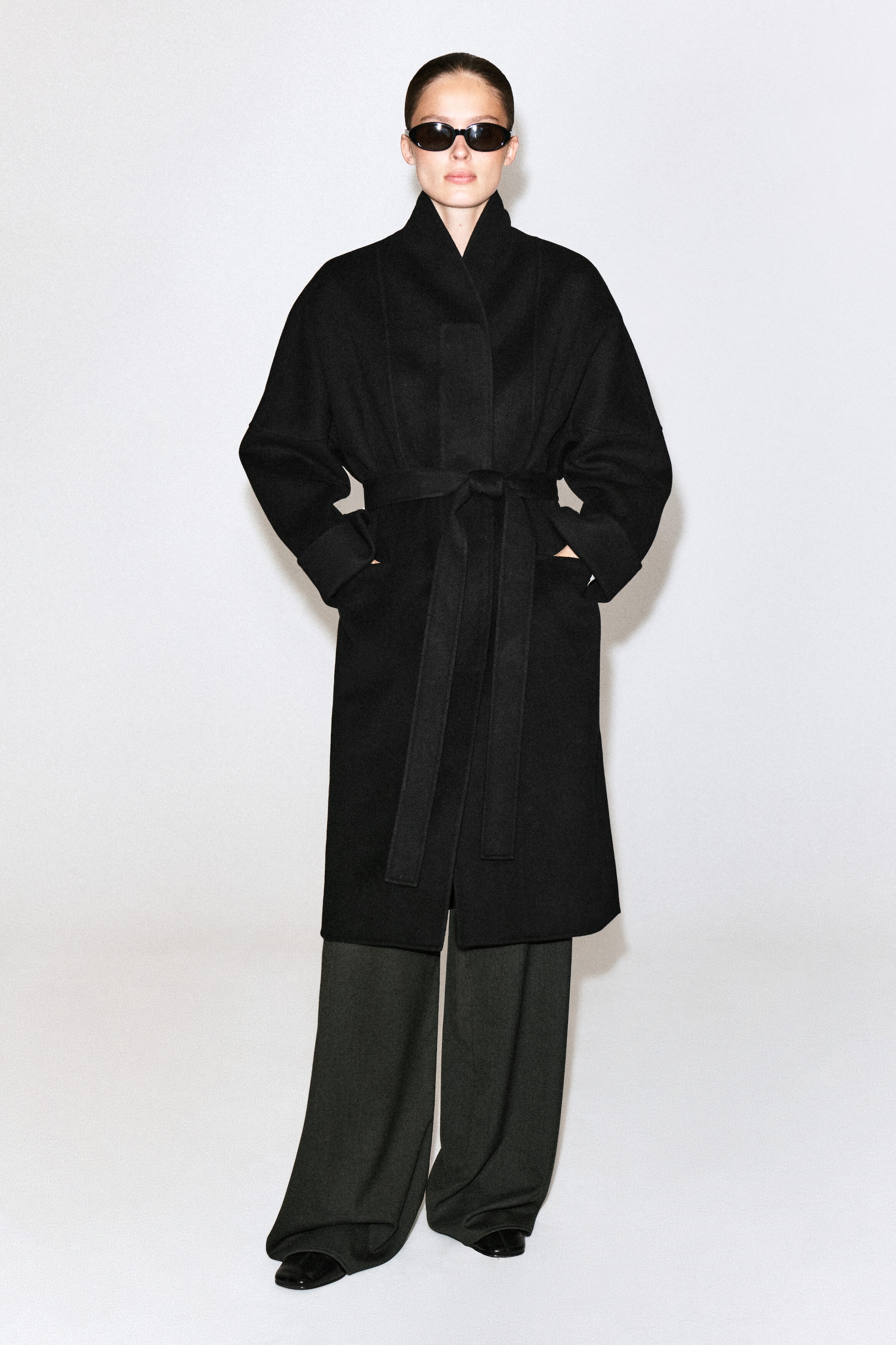 Un manteau-shawl oversize en laine double face noir est porté par le mannequin, avec un col montant, des épaules tombantes et une ceinture à nouer à la taille. Le mannequin est debout, les mains glissées dans les poches du manteau, et le porte avec un pantalon large foncé. Un manteau-shawl oversize en laine double face noir est porté par le mannequin, avec un col montant, des épaules tombantes et une ceinture à nouer à la taille. Le mannequin est debout, les mains glissées dans les poches du manteau, et le porte avec un pantalon large foncé.