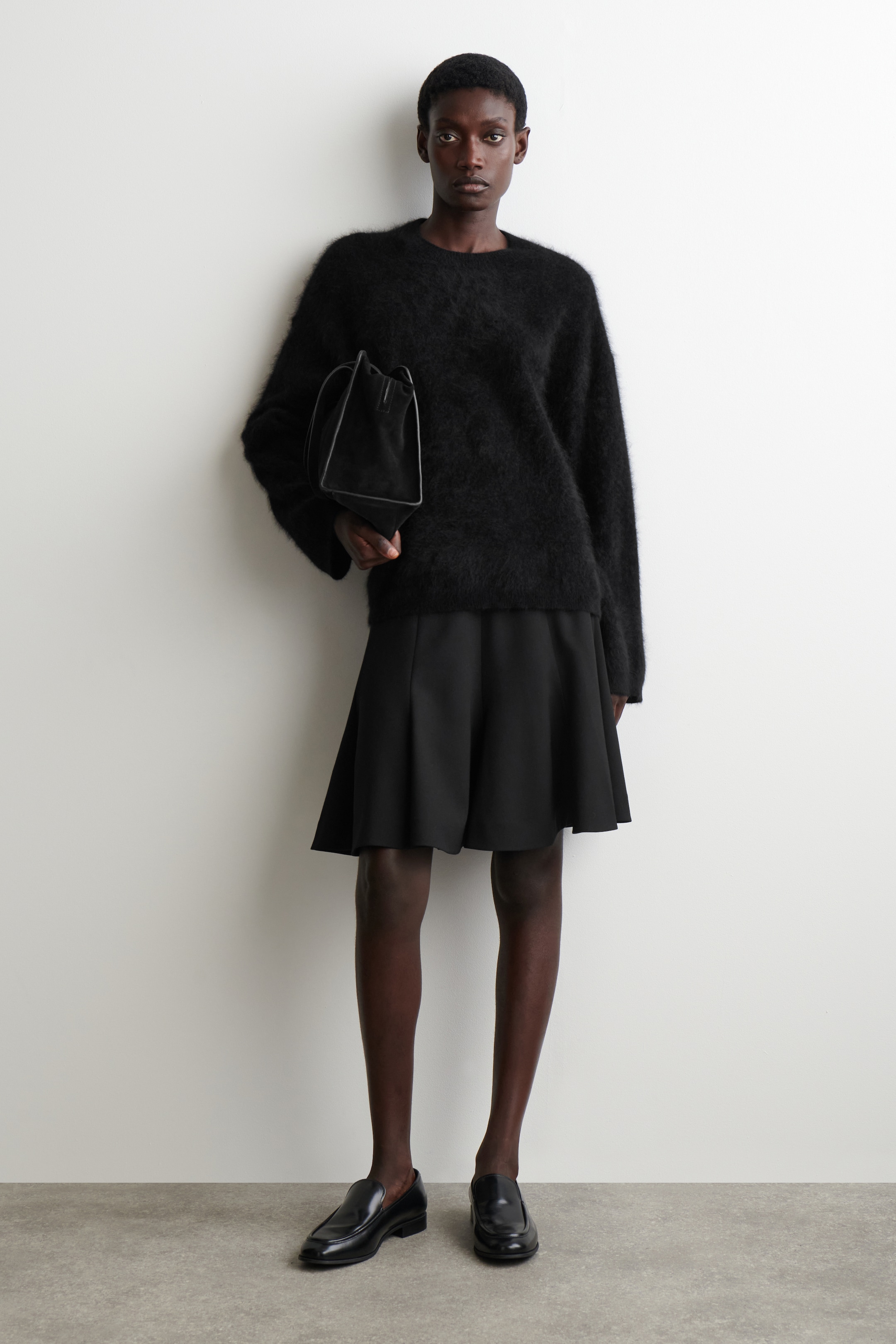 Se større billede: En sort, fuzzy crew-neck sweater med en oversized pasform præsenteres på en fuld-længde figur, stylet med sorte A-line culotte shorts. En lille sort struktureret håndtaske holdes, og sorte polerede slip-on loafers bæres.