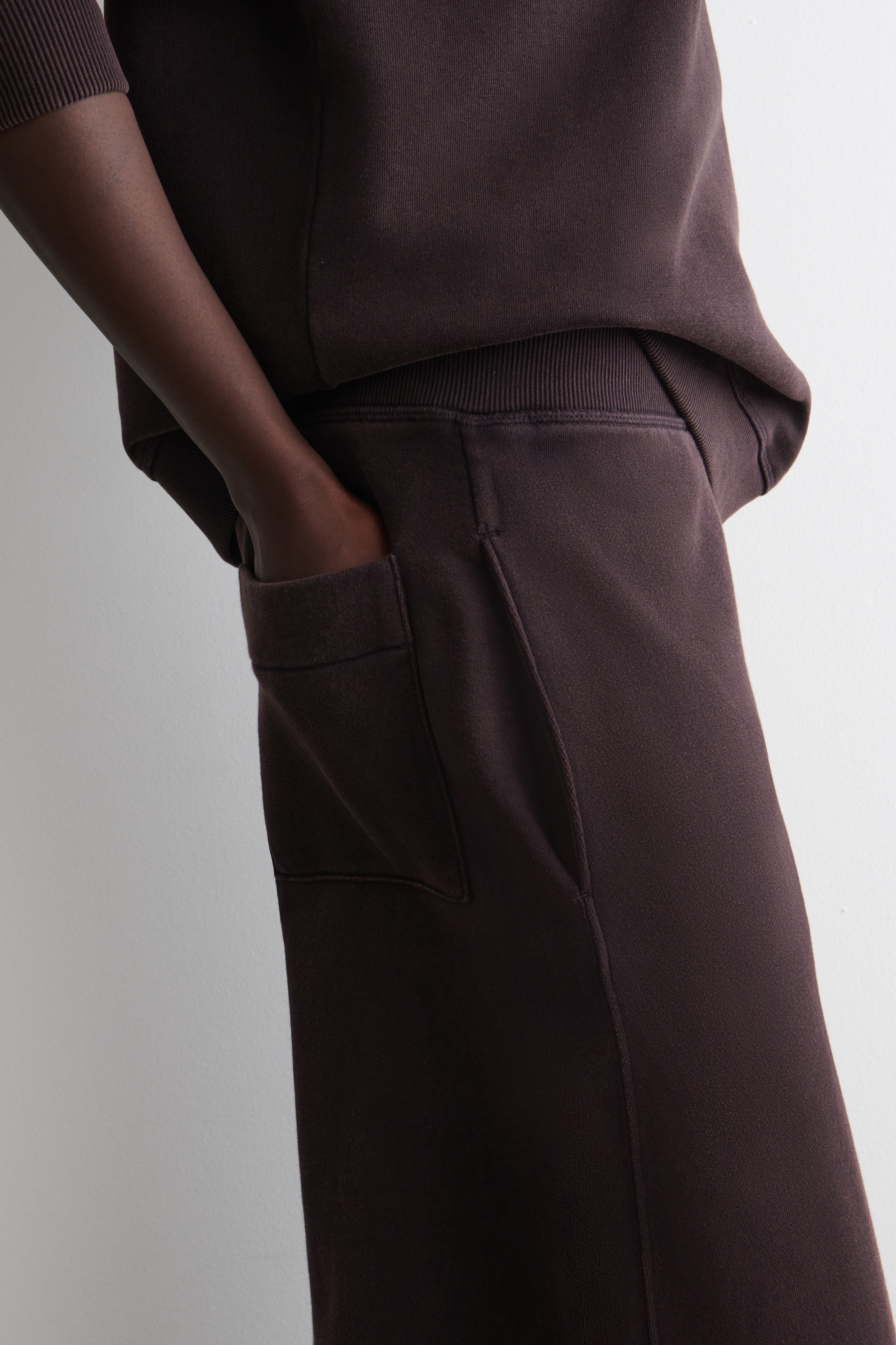 Ingrandisci l'immagine: I joggers marrone scuro sono indossati, angolati a DESTRA, con una cintura elastica a coste. Una mano è appoggiata in una tasca laterale, con una tasca a toppa rettangolare visibile sotto di essa sulla parte superiore della gamba, e sono abbinati a una felpa marrone scuro coordinata.