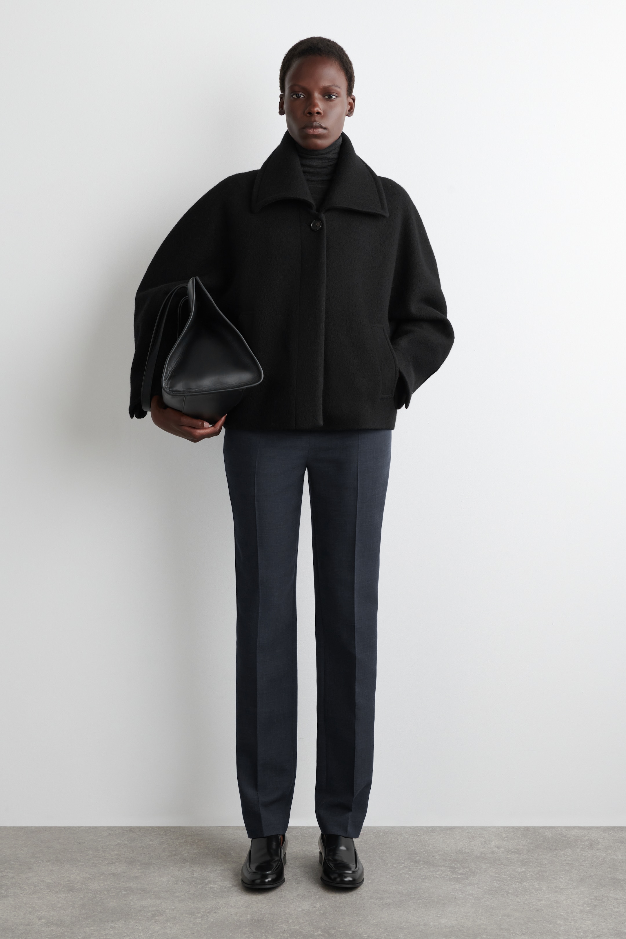 Grösseres Bild ansehen: Ein komplettes Outfit wird präsentiert, das eine schwarze, kurze, nach vorne gerichtete Jacke aus gebürstetem Stoff mit breitem Kragen und einem Knopfverschluss sowie Seitentaschen umfasst. Es wird über einem dunklen, gerippten Rollkragenpullover gestylt und mit dunklen, gerade geschnittenen, maßgeschneiderten Hosen und schwarzen Anzugschuhen kombiniert. Eine schwarze, strukturierte Ledertasche wird in der Hand gehalten.