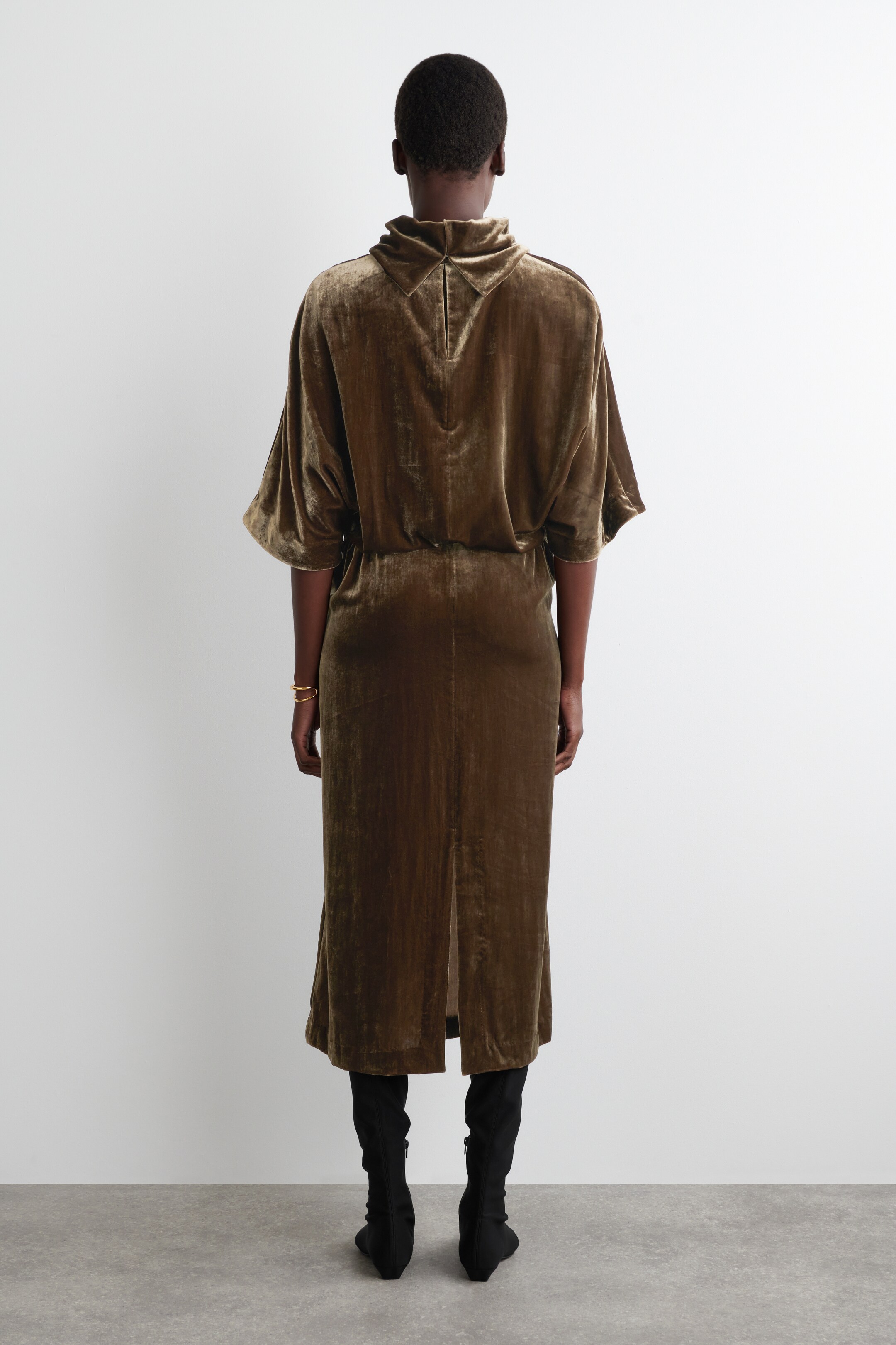 Grösseres Bild ansehen: Ein olivbraunes Samtmidikleid wird von hinten gezeigt, mit einem hohen Kragen und einem V-förmigen Ausschnitt am Nacken sowie weiten, ellbogenlangen Dolman-Ärmeln. Das Kleid wird an der Taille gerafft, fließt in einen geraden Rock mit Rückenschlitz und wird mit schwarzen Stiefeln getragen.