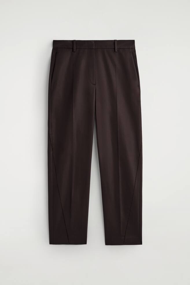 Vista FRONTAL de pantalones marrón oscuro, con una cintura plana con trabillas para cinturón y bolsillos en las costuras laterales. Pliegues marcados recorren la parte delantera de cada pierna, y un delicado bordado abierto detalla una línea diagonal en el lado exterior de cada pierna, extendiéndose desde la parte superior del muslo hasta el dobladillo. - 1