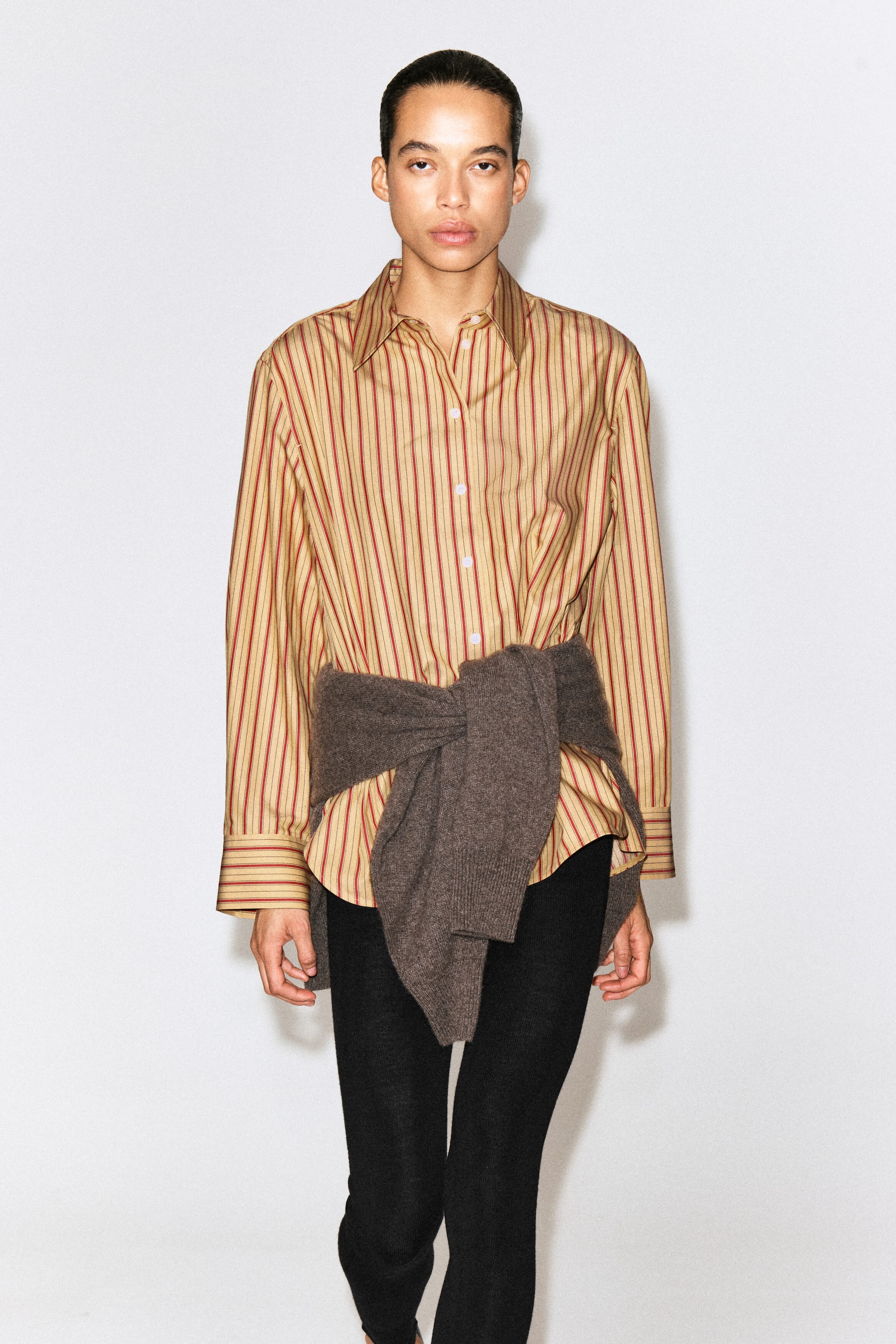 The striped cotton-lyocell shirt is shown from the front, featuring vertical red and brown stripes on a beige base, a pointed collar, and long sleeves with buttoned cuffs. A brown knit sweater is worn tied around the waist, partially layering over the shirt’s relaxed fit.