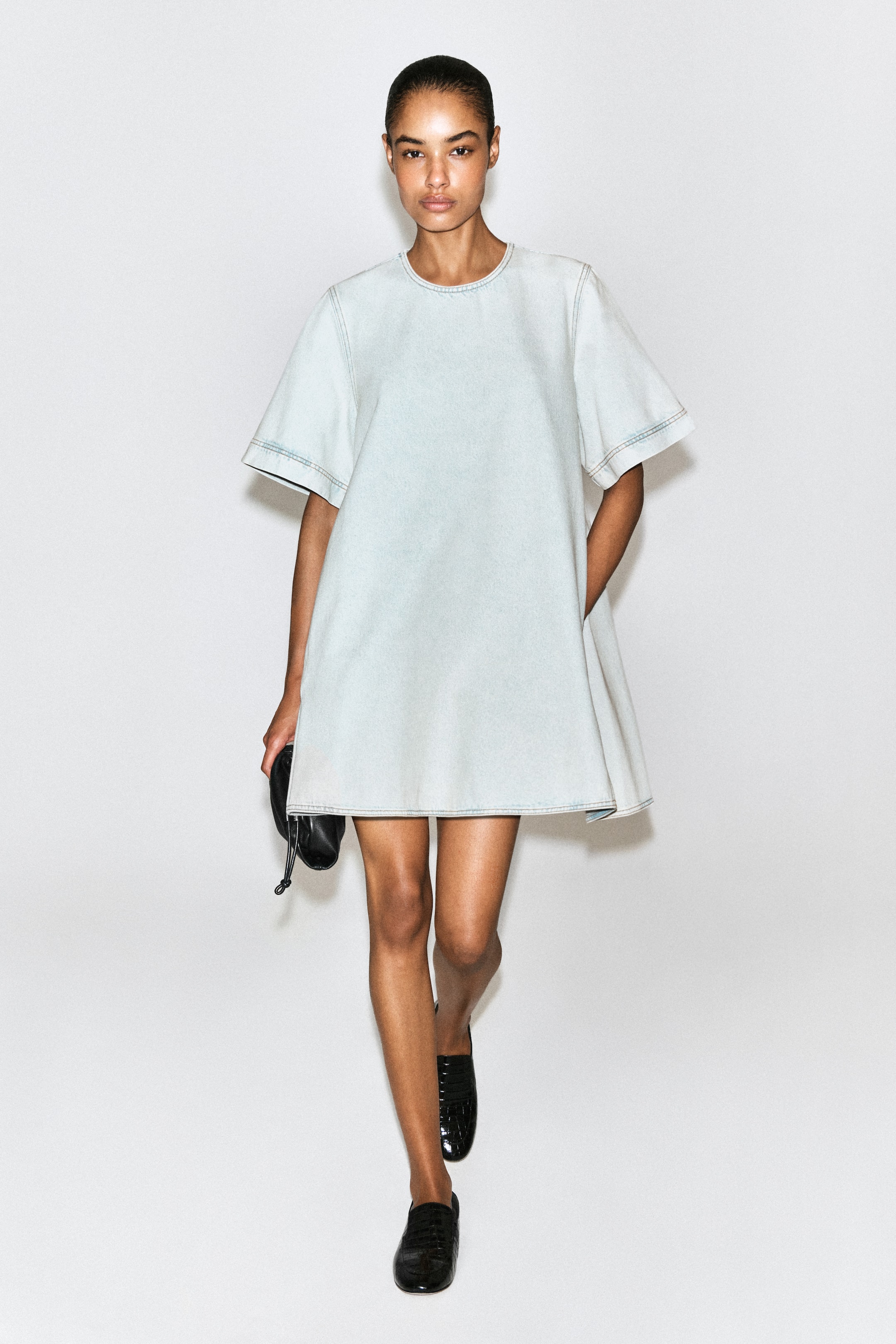 Ver imagen más grande: Un minivestido de denim azul claro presenta un escote redondo, mangas cortas y anchas, pespuntes en contraste y discretos bolsillos laterales, mostrando una silueta relajada en línea A desde el frente.