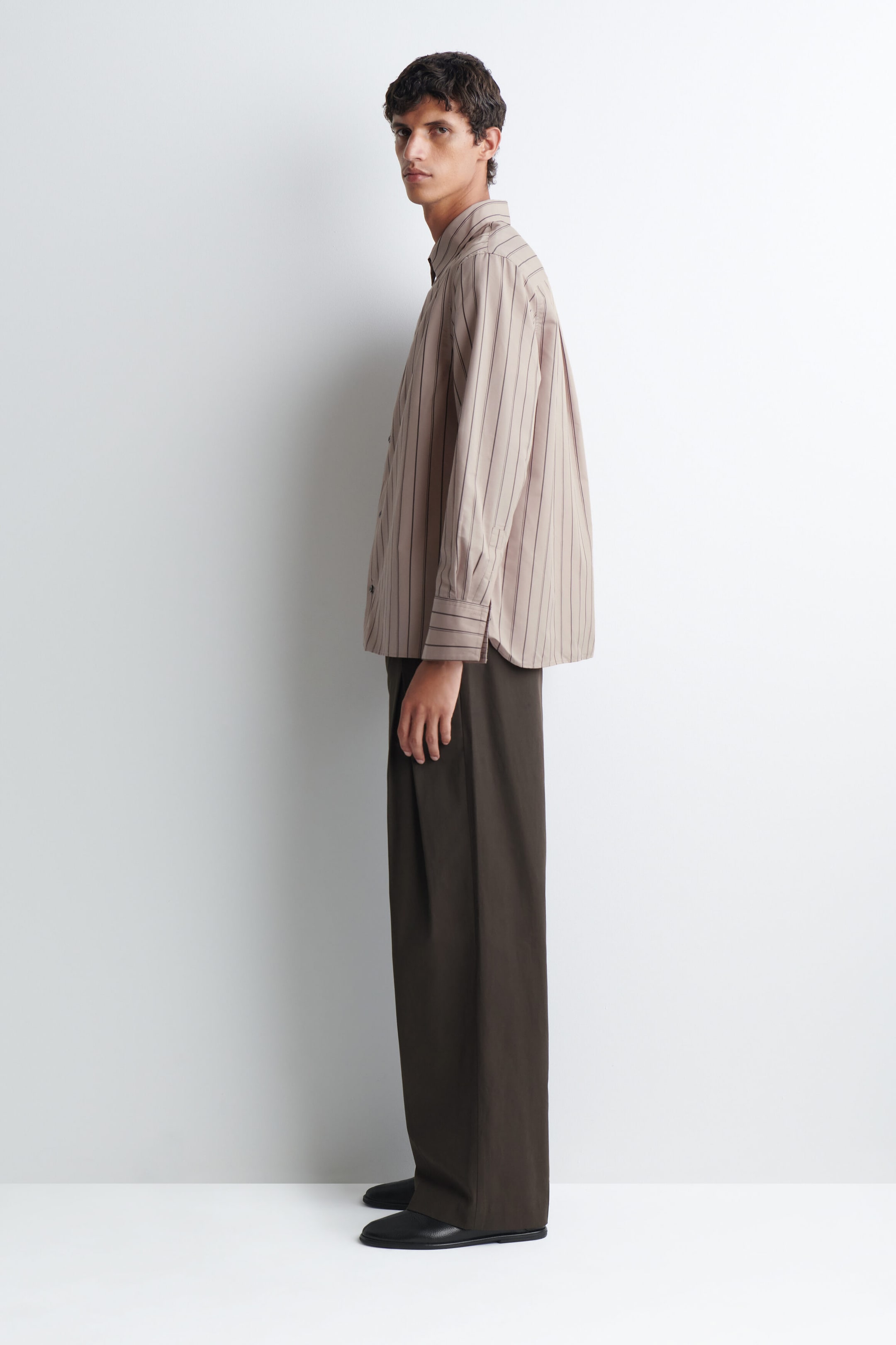 The striped structured cotton-silk shirt is worn by the model, featuring a light brown fabric with thin, dark vertical stripes, a point collar and wide cuffs. The relaxed-fit shirt is styled untucked and paired with dark brown wide-leg trousers. The striped structured cotton-silk shirt is worn by the model, featuring a light brown fabric with thin, dark vertical stripes, a point collar and wide cuffs. The relaxed-fit shirt is styled untucked and paired with dark brown wide-leg trousers.