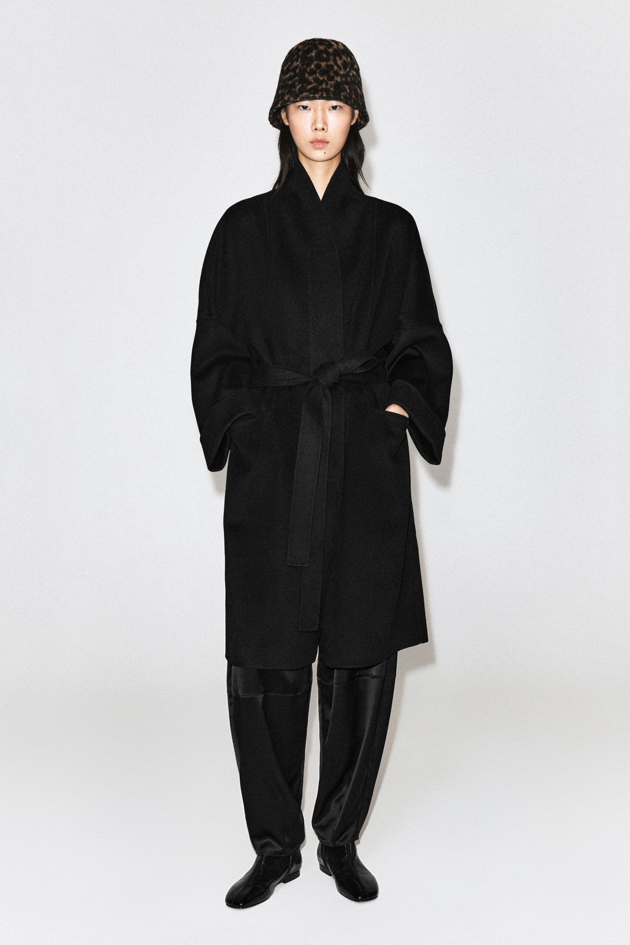 A leopard-print bucket hat with a textured, boiled wool finish and a gently curved brim is being worn by the model. The hat is styled with a long, belted black coat and matching black trousers. A leopard-print bucket hat with a textured, boiled wool finish and a gently curved brim is being worn by the model. The hat is styled with a long, belted black coat and matching black trousers.