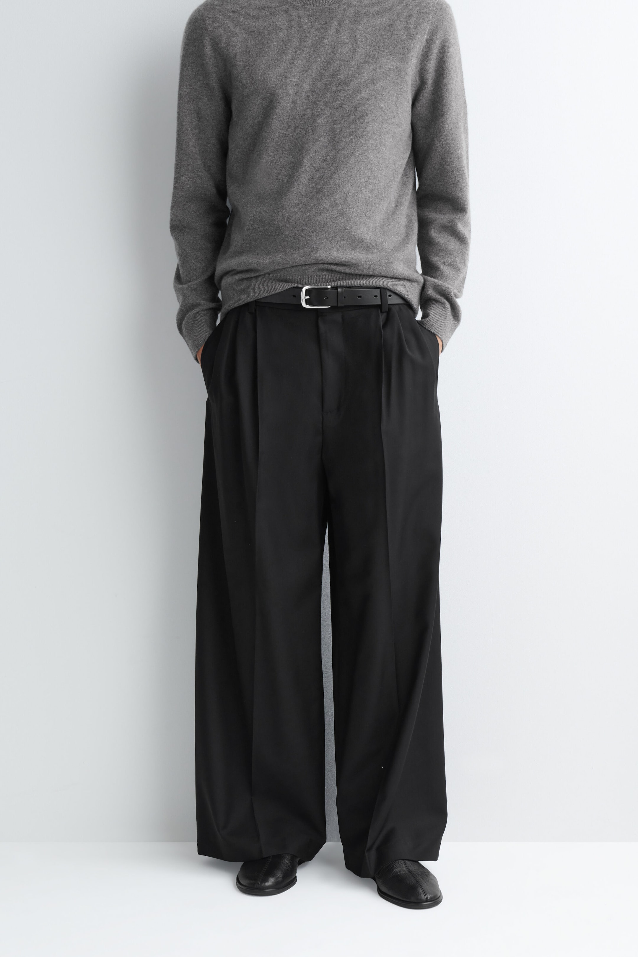 Die schwarzfarbene, weit geschnittene Hose mit legerer Passform und Bügelfalten wird vom Model getragen, das mit den Händen in den Taschen steht. Die Hose verfügt über doppelte Frontfalten und einen entspannten Fall und ist mit einem schwarzen Ledergürtel, einem grauen Langarm-Pullover und schwarzen Lederschuhen kombiniert.