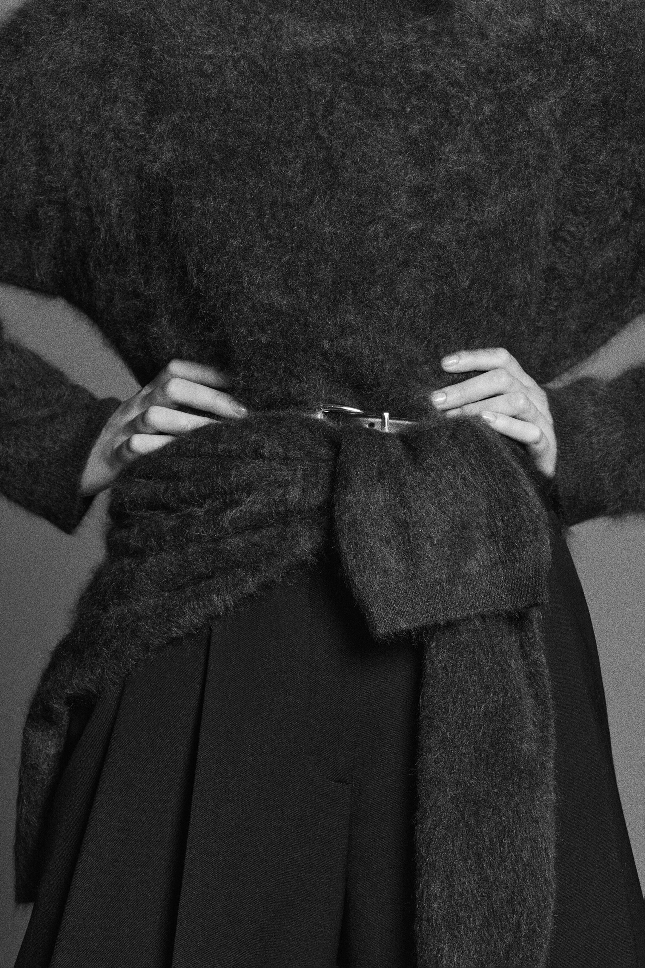 A front view of a dark, heavily textured, brushed knit jumper styled with a second matching jumper tied around the waist and secured with a thin belt. The knitwear features a fuzzy, tactile surface and is paired with dark pleated trousers.