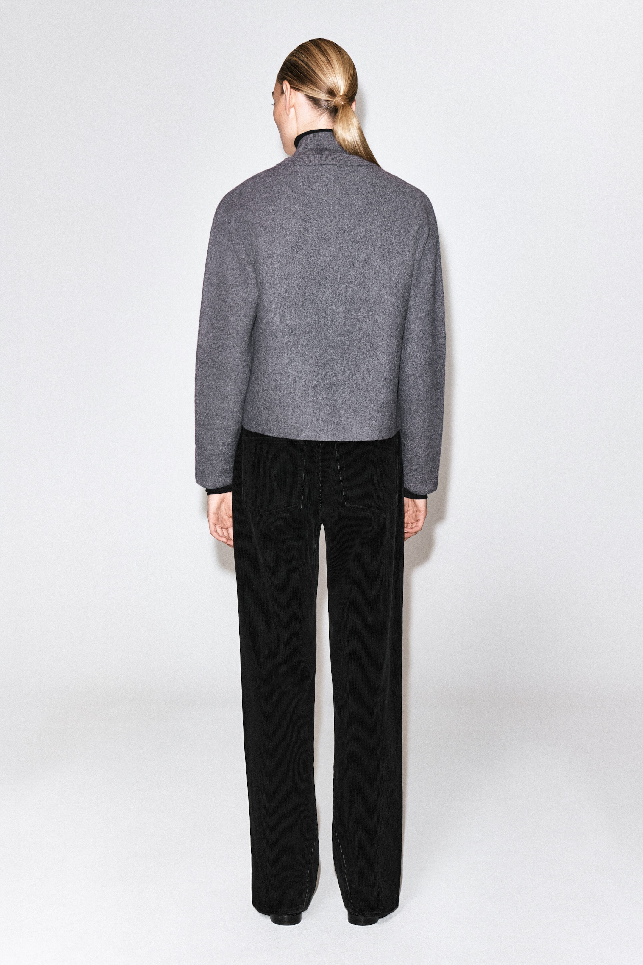A grey boiled wool cardigan with a rounded silhouette is being worn by the model, who is standing with their back to the camera. The cardigan features a high collar and cuffs with visible black under-layering, and is paired with black trousers. A grey boiled wool cardigan with a rounded silhouette is being worn by the model, who is standing with their back to the camera. The cardigan features a high collar and cuffs with visible black under-layering, and is paired with black trousers.