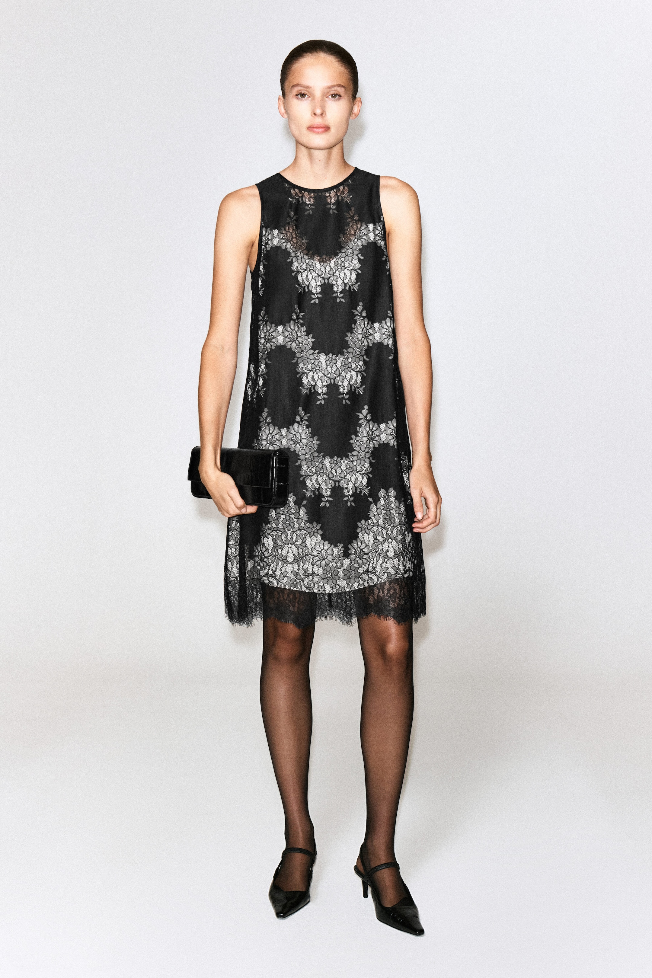 The model is wearing a sleeveless lace midi dress, featuring a black floral lace overlay with a contrasting light-coloured pattern underneath and a scalloped eyelash lace hem. The outfit is paired with sheer black tights, black pointed-toe heels, and a black rectangular clutch bag held by the model. The model is wearing a sleeveless lace midi dress, featuring a black floral lace overlay with a contrasting light-coloured pattern underneath and a scalloped eyelash lace hem. The outfit is paired with sheer black tights, black pointed-toe heels, and a black rectangular clutch bag held by the model.