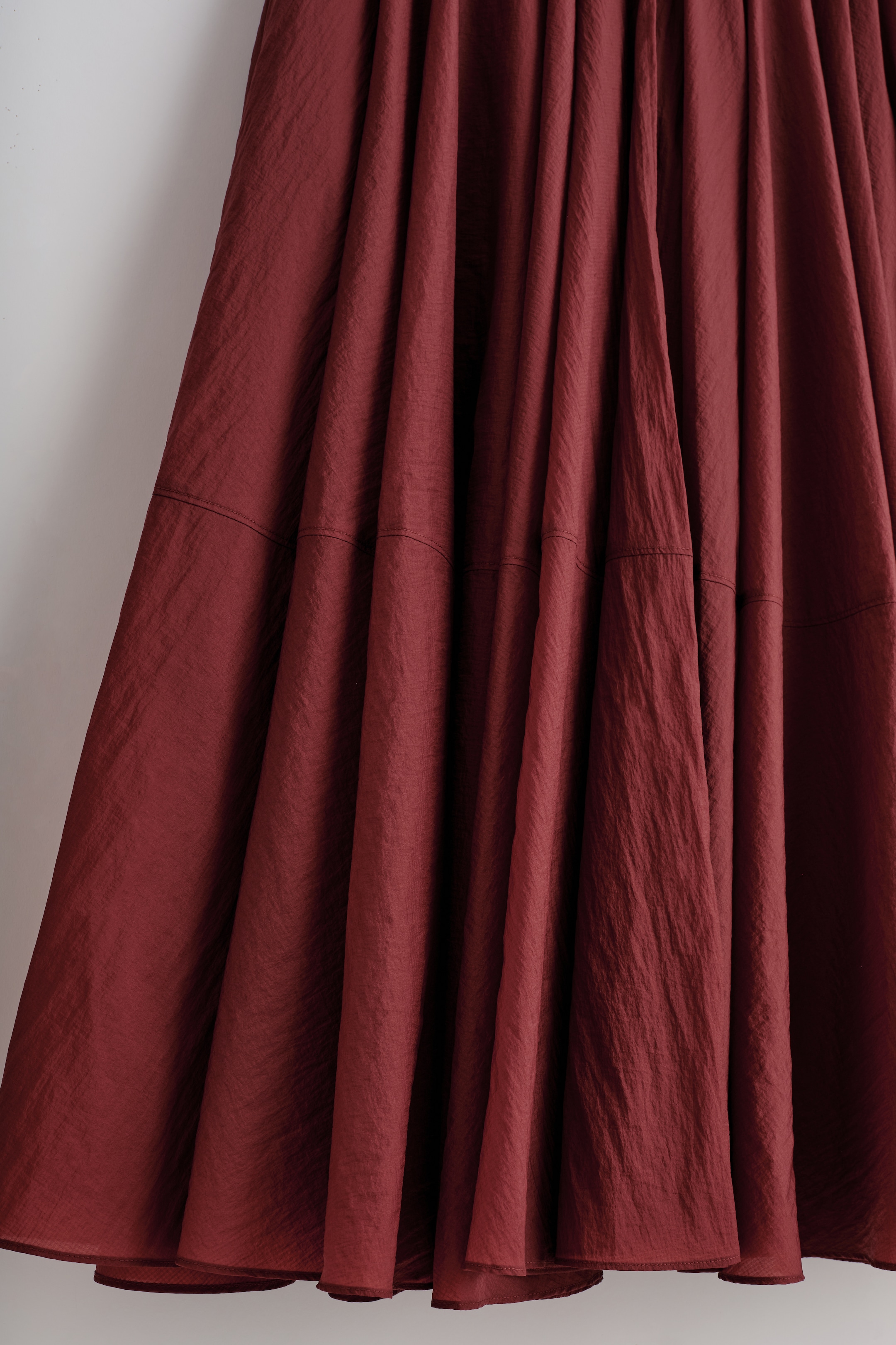 Agrandir l'image: Le devant d'une jupe rouge foncé présente une silhouette volumineuse et froncée avec une texture froissée. Des lignes de couture horizontales sont visibles sur la partie inférieure du tissu, contribuant à son drapé ample.