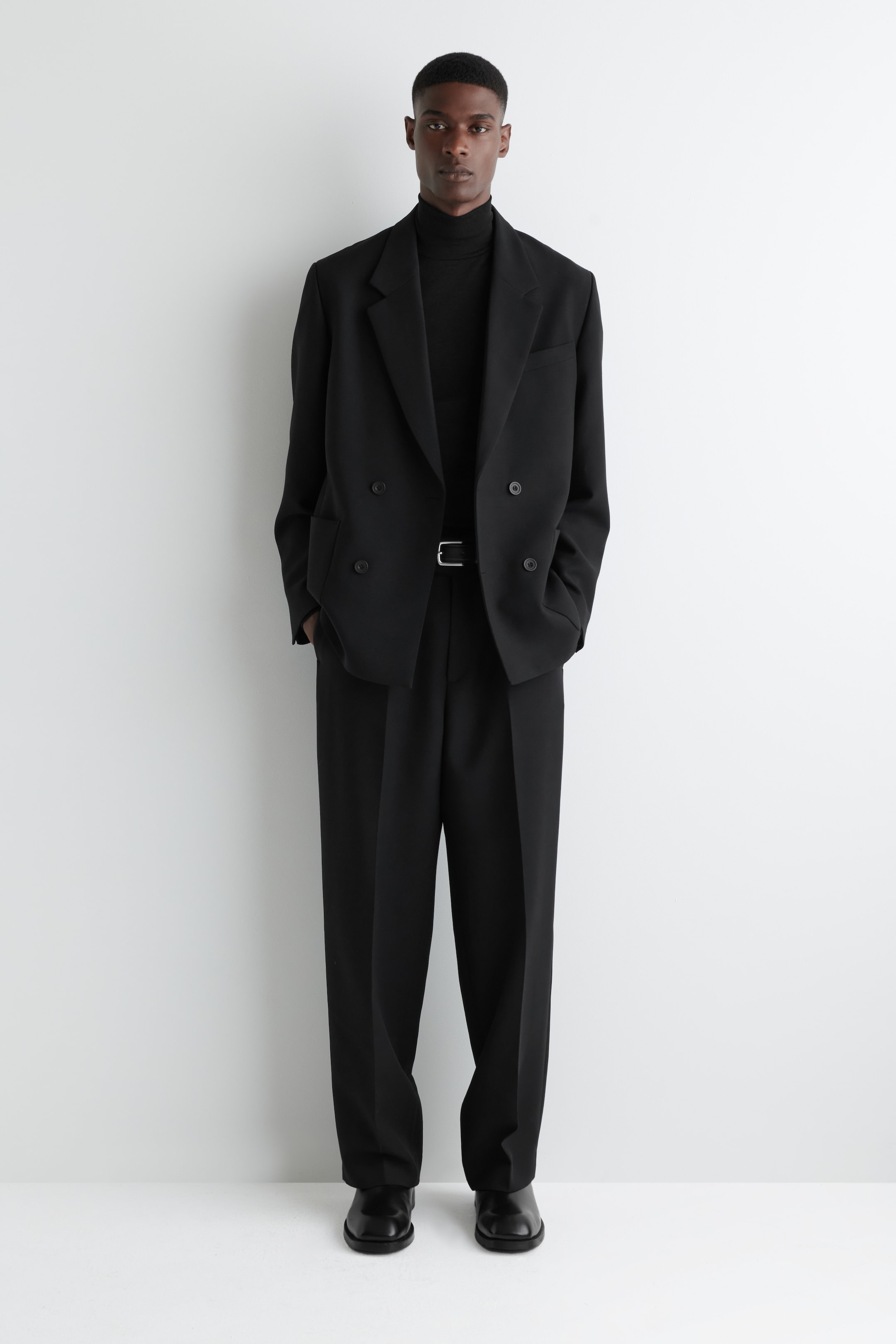 Agrandir l'image: Un costume noir est présenté de face, composé d'un blazer croisé à revers crantés et quatre boutons visibles, et d'un pantalon large assorti. Un pull à col roulé noir est porté sous le blazer, complété par une ceinture noire avec une boucle argentée et des chaussures formelles noires.