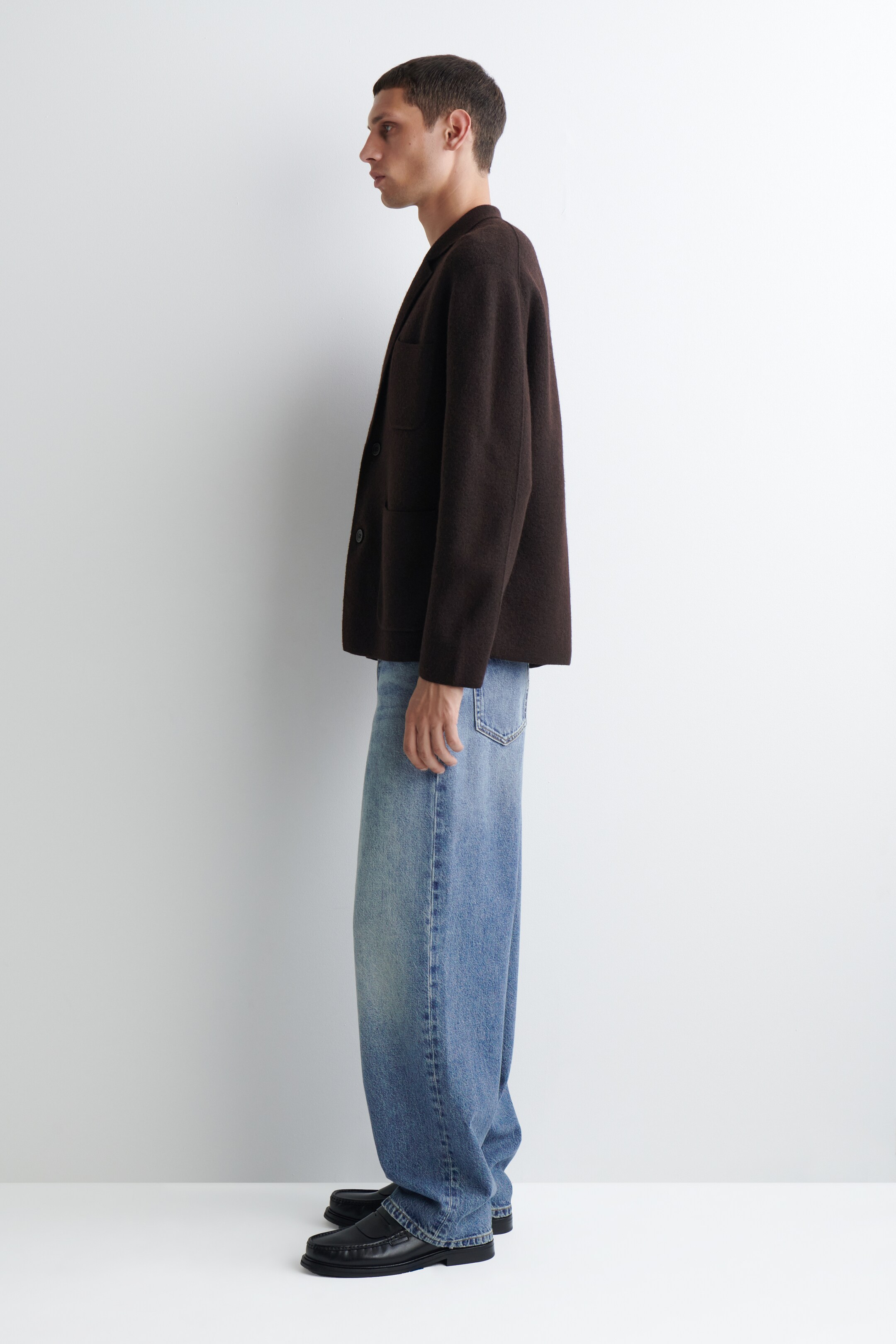 Ver imagem maior: Um blazer de feltro castanho escuro, com lapelas de entalhe e dois botões visíveis, é apresentado de perfil lateral. É combinado com jeans de perna larga azul claro e mocassins pretos, exibindo uma silhueta descontraída.