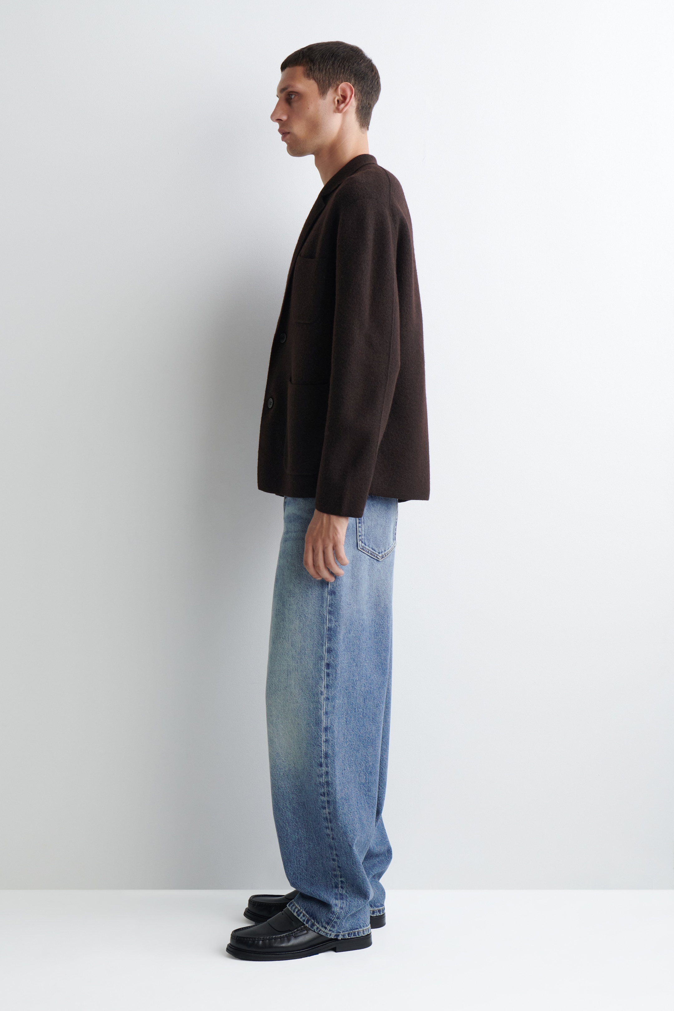 Größeres Bild ansehen: Ein dunkelbrauner, gefilzter Blazer mit Kerbrevers und zwei sichtbaren Knöpfen wird im Seitenprofil präsentiert. Er wird mit hellblauen, weit geschnittenen Jeans und schwarzen Loafers gestylt, was eine entspannte Silhouette zeigt.