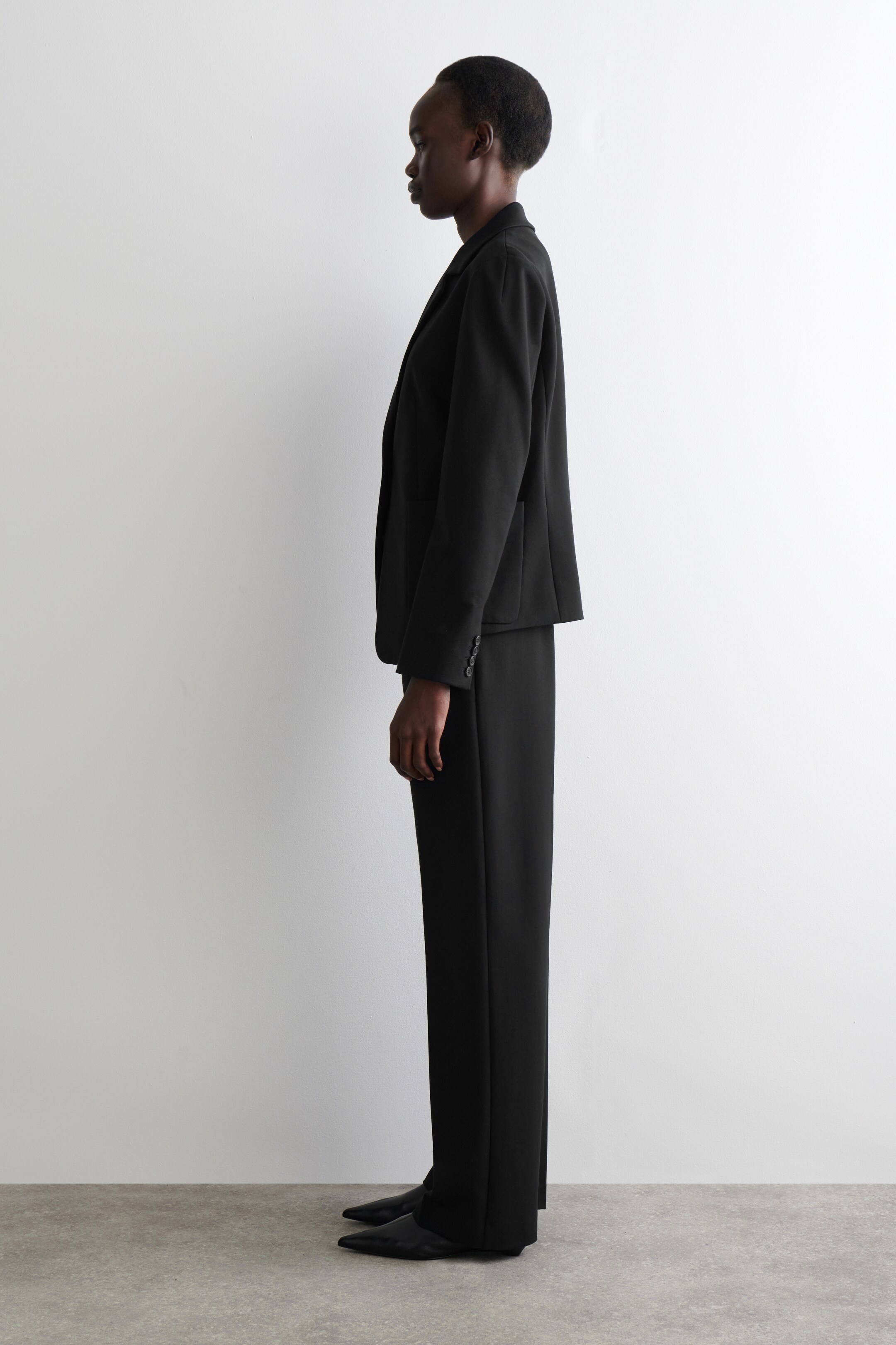 Zobrazit větší obrázek: Černý dvoudílný oblek je zobrazen z bočního profilu, obsahuje strukturované sako s dlouhými rukávy, třemi knoflíky na manžetách a viditelnými bočními kapsami, doplněný kalhotami s rovnými nohavicemi.