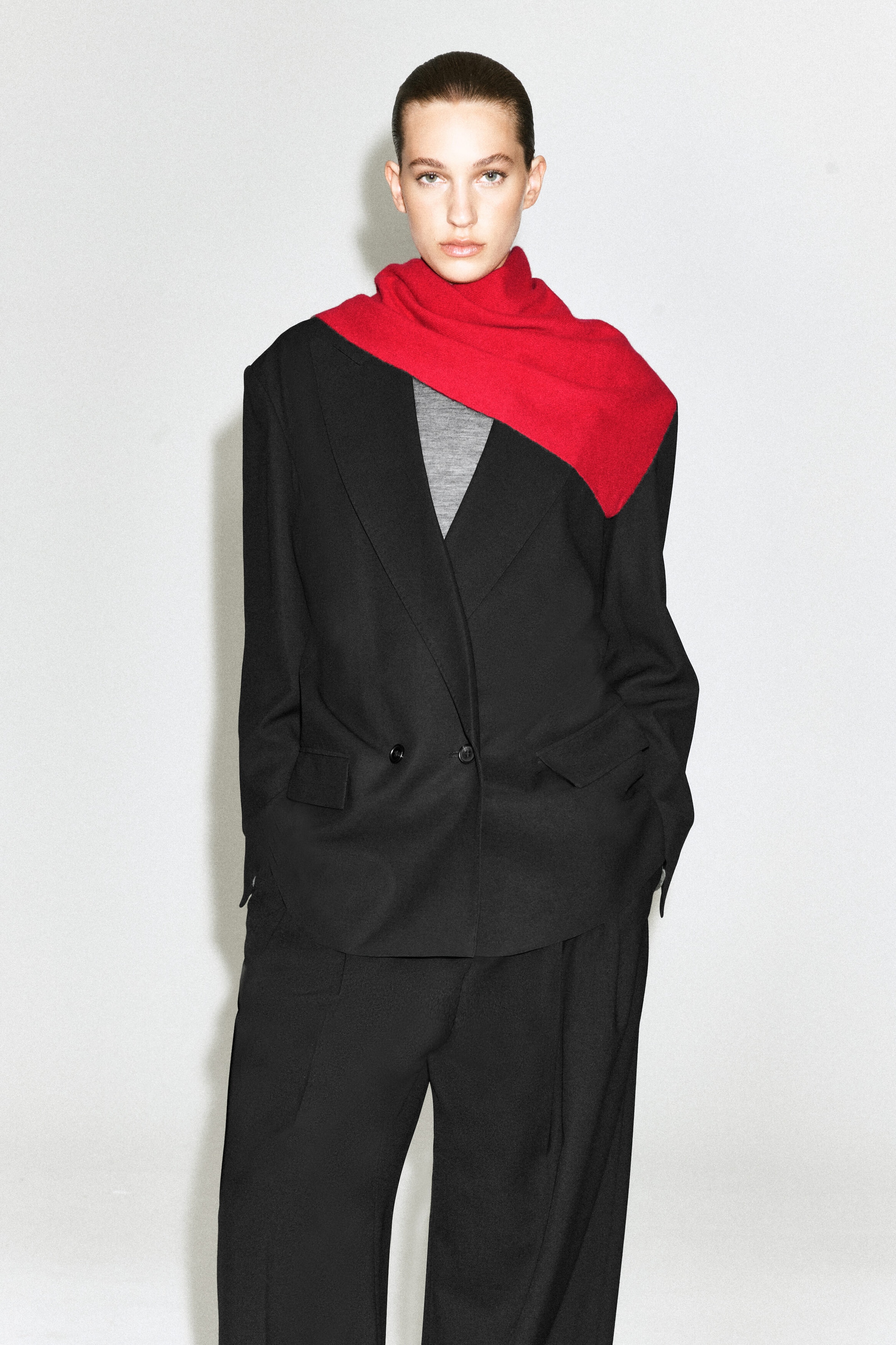 A red cashmere triangle scarf is worn wrapped around the model’s neck, with one side draped asymmetrically over the shoulder of a black double-breasted blazer. The vibrant knit scarf is styled over the blazer and a gray top, creating a layered look. A red cashmere triangle scarf is worn wrapped around the model’s neck, with one side draped asymmetrically over the shoulder of a black double-breasted blazer. The vibrant knit scarf is styled over the blazer and a gray top, creating a layered look.