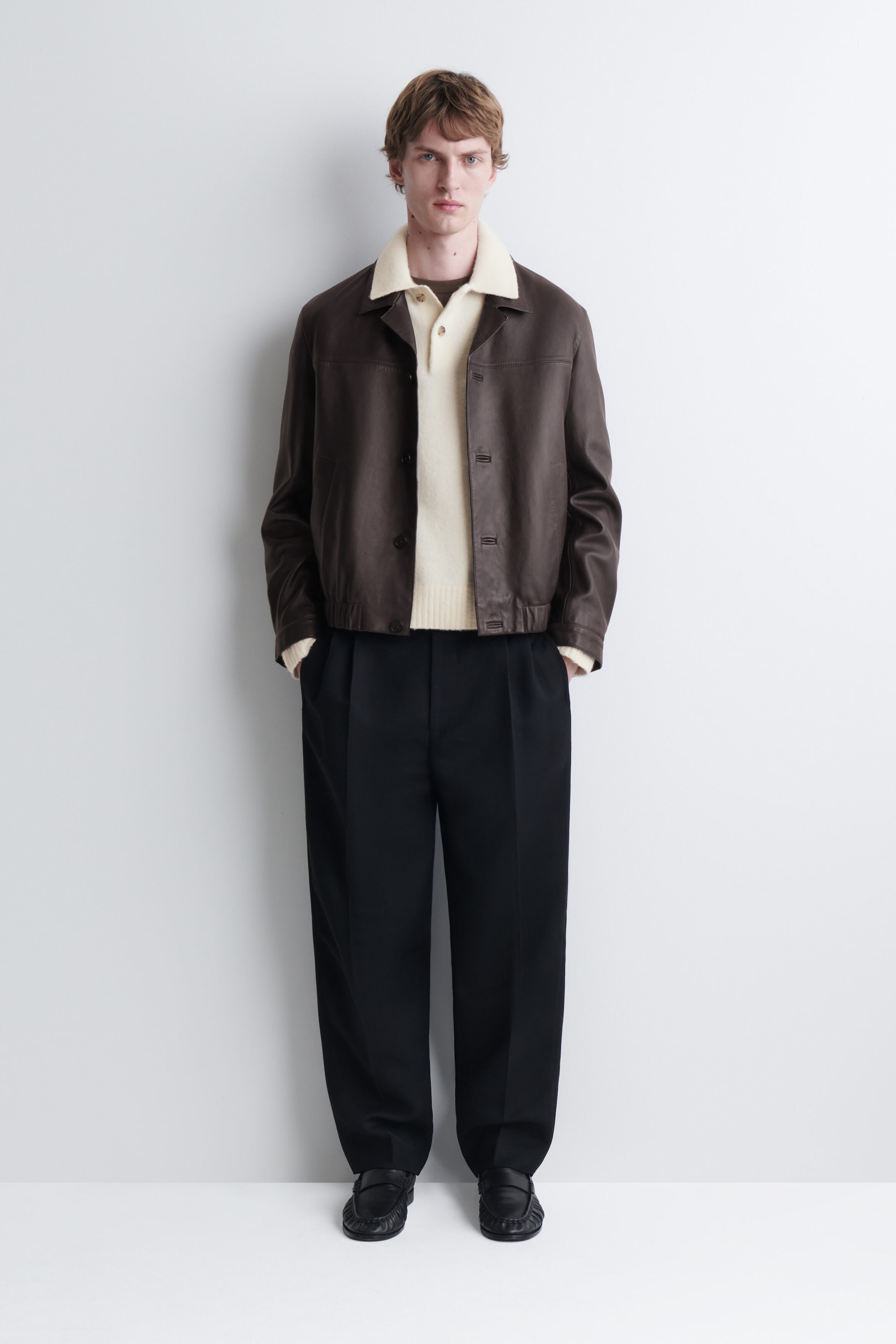 A cream-colored brushed wool polo shirt featuring a classic collar and a button placket is being worn by the model. The shirt is layered under an open dark brown leather jacket and paired with black wide-leg trousers.