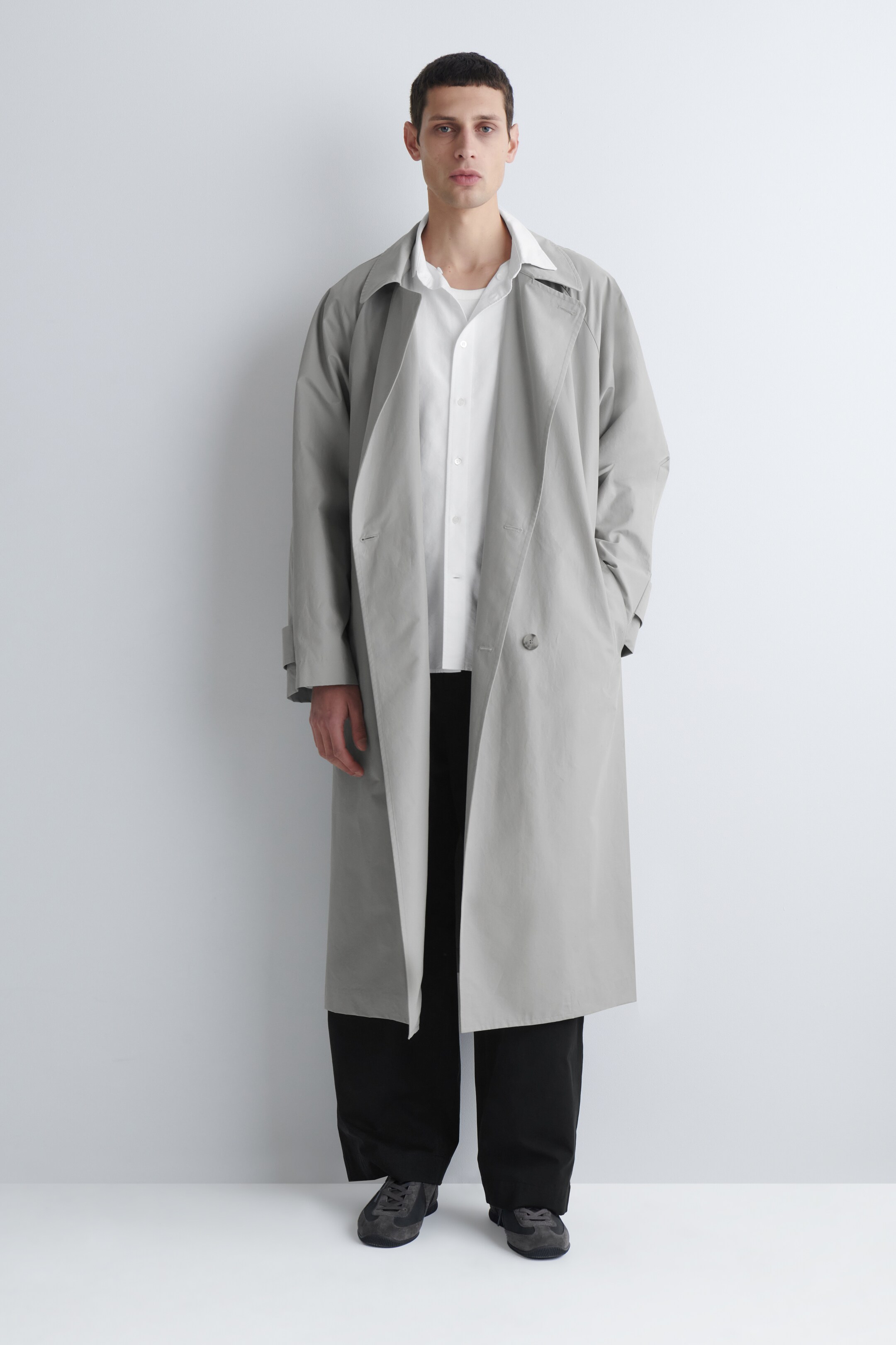 Größeres Bild ansehen: Ein hellgrauer Trenchcoat wird VON VORNE gezeigt und zeichnet sich durch eine entspannte, lange Silhouette aus, die unterhalb der Knie endet. Er wird offen über einem weißen Hemd mit Kragen gestylt, wodurch sein breiter Kragen und zwei sichtbare Knöpfe an der ungeknöpften doppelreihigen Vorderseite zur Geltung kommen.
