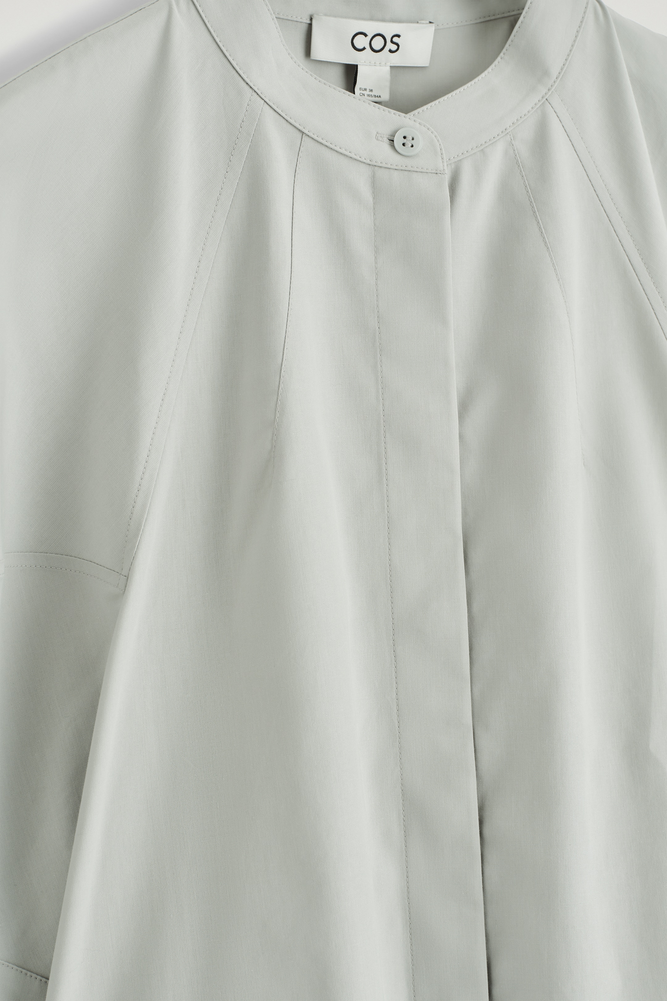 Grösseres Bild ansehen: Eine hellgraue Bluse wird von vorne präsentiert, mit einem abgerundeten Großvaterkragen und einem sichtbaren Knopfverschluss am Halsausschnitt sowie einer verdeckten Knopfleiste. Raglanärmel sind durch diagonale Nahtlinien gekennzeichnet, die sich vom Kragen aus erstrecken, und der Stoff fällt glatt.