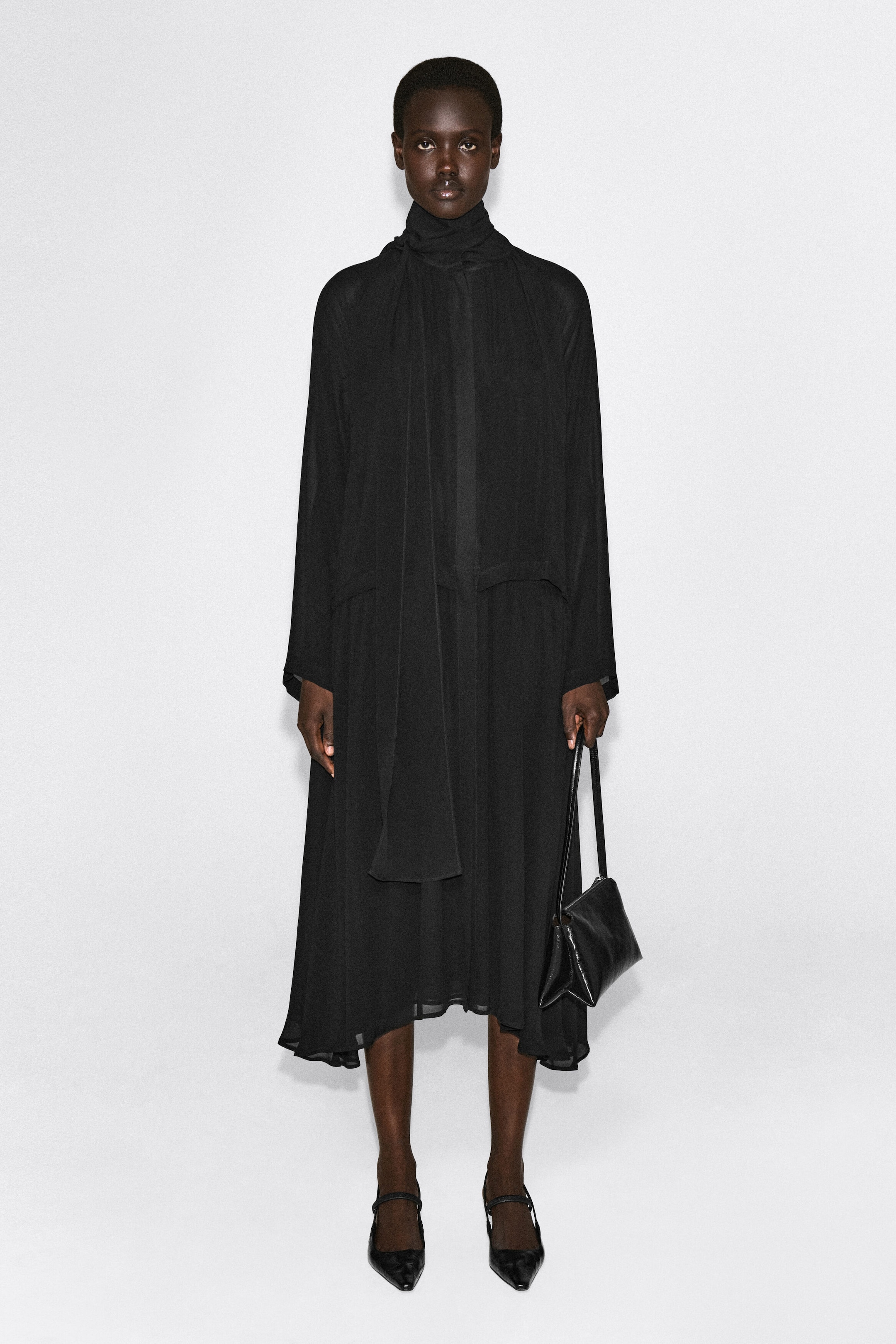 Shown from the front, the black fluid scarf-detail midi dress features a relaxed, loose-fitting silhouette with long sleeves and a high neckline. A long, integrated scarf drapes down the center front of the dress, which falls to a midi length with a slightly asymmetrical hem.