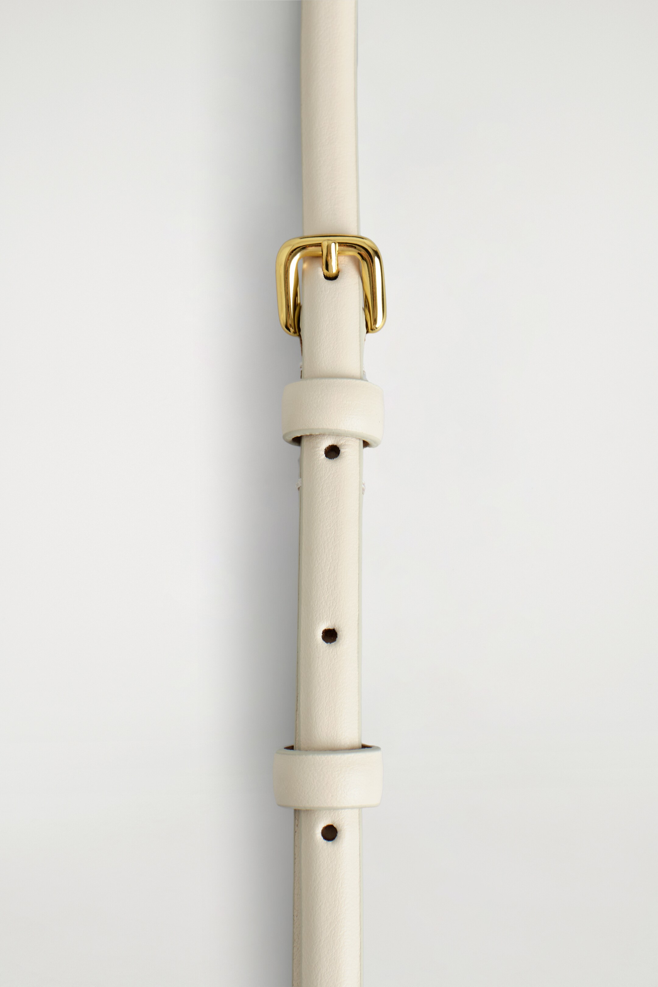 Zobrazit větší obrázek: Úzký, krémově zbarvený kožený pásek je položen vertikálně, s klasickou zlatou sponou na vrcholu. Pásek má několik kulatých nastavitelných otvorů a dva odpovídající kožené poutka.